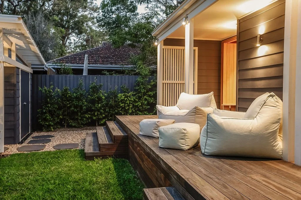 Outdoor patio with wooden decking, beige bean bag chairs, and a small SUP board, at twilight with exterior lights on