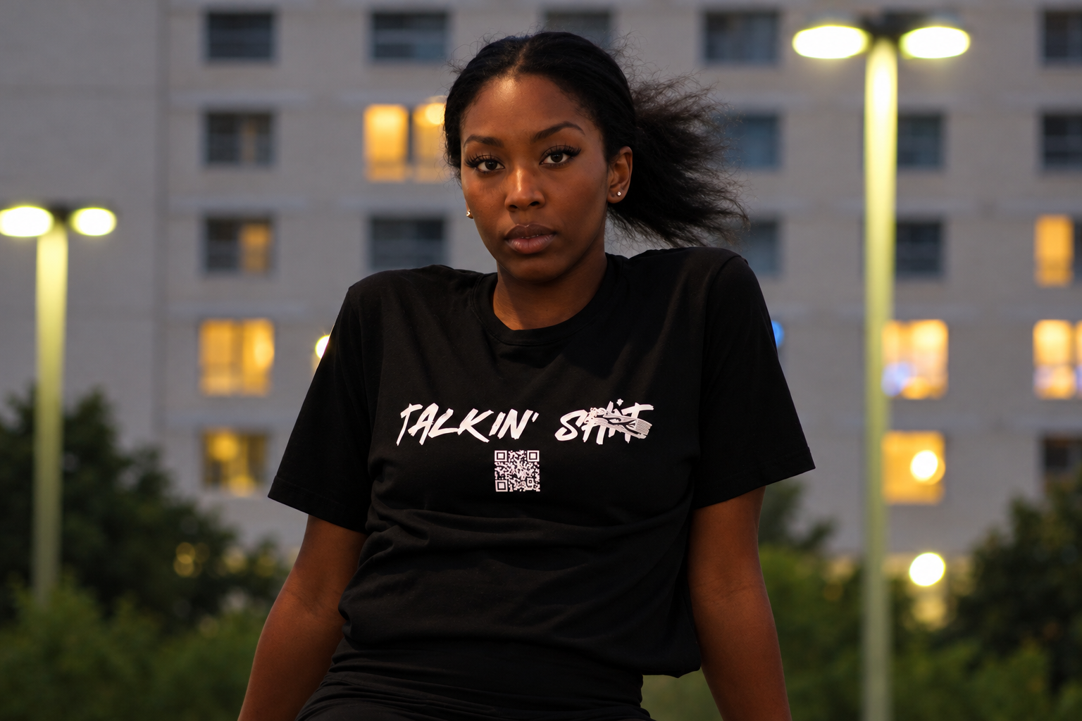 Young Black woman wearing a black “Talkin’ SICK” graphic T-shirt with a QR code outdoors at dusk.