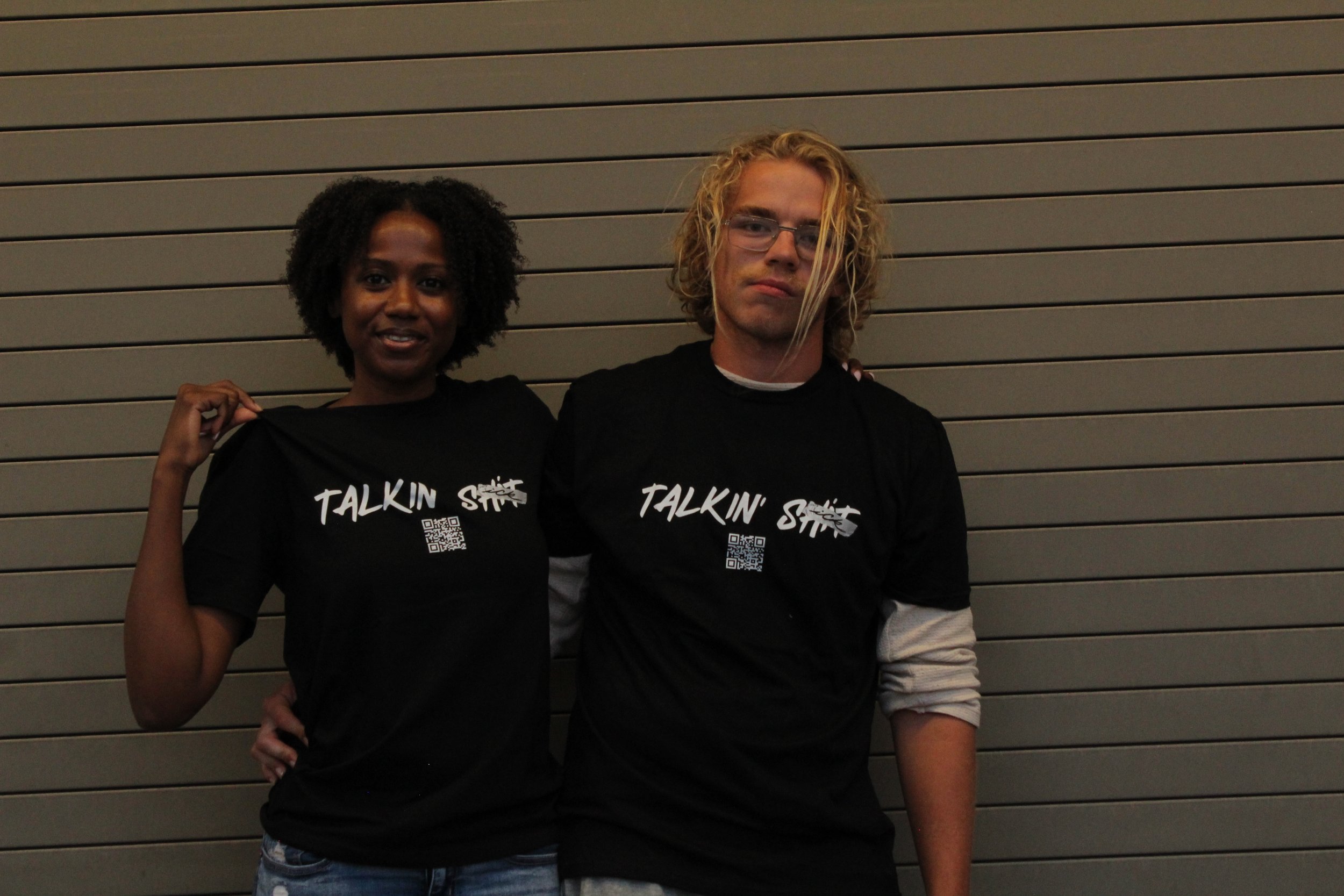 Two people wearing matching black “Talkin’ SICK” T-shirts pose together in front of a paneled wall.