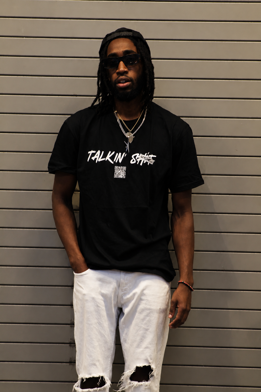 Man with dreadlocks and sunglasses wearing a black graphic T-shirt, white ripped shorts, layered necklaces, and a backward cap in front of a metal shutter. wearing a black “Talkin’ SICK” graphic T-shirt with a QR code
