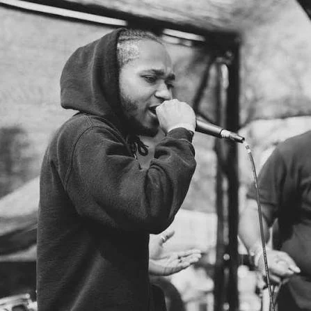 Black-and-white photo of Qu33zy rapping into a microphone at an outdoor performance.