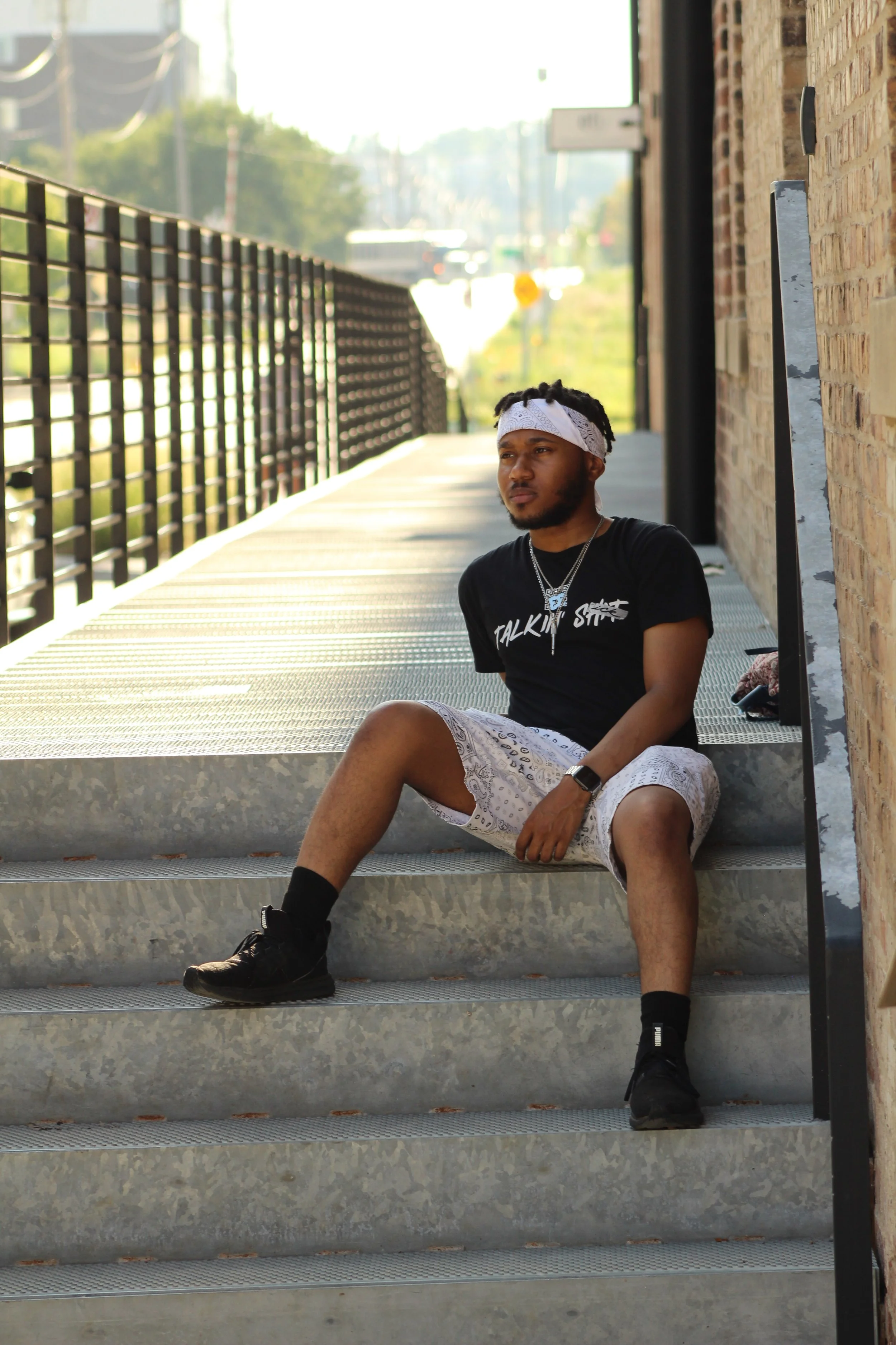 Qu33zy wearing a white bandana and wearing a black “Talkin’ SICK” graphic T-shirt with a QR code sitting on metal steps beside a brick building and railing