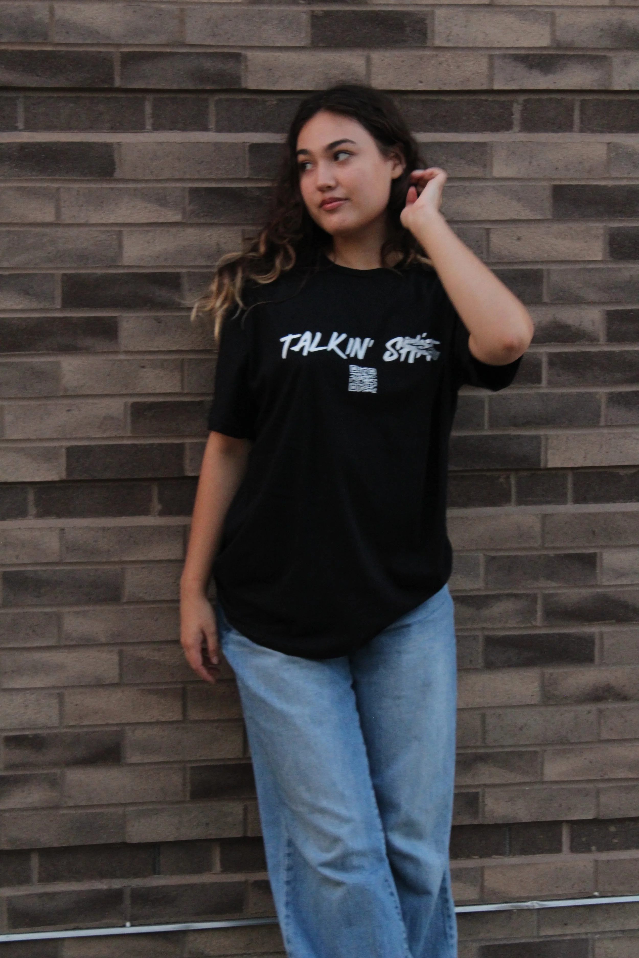 Woman in a black graphic T-shirt and jeans posing against a brick wall. wearing a black “Talkin’ SICK” graphic T-shirt with a QR code