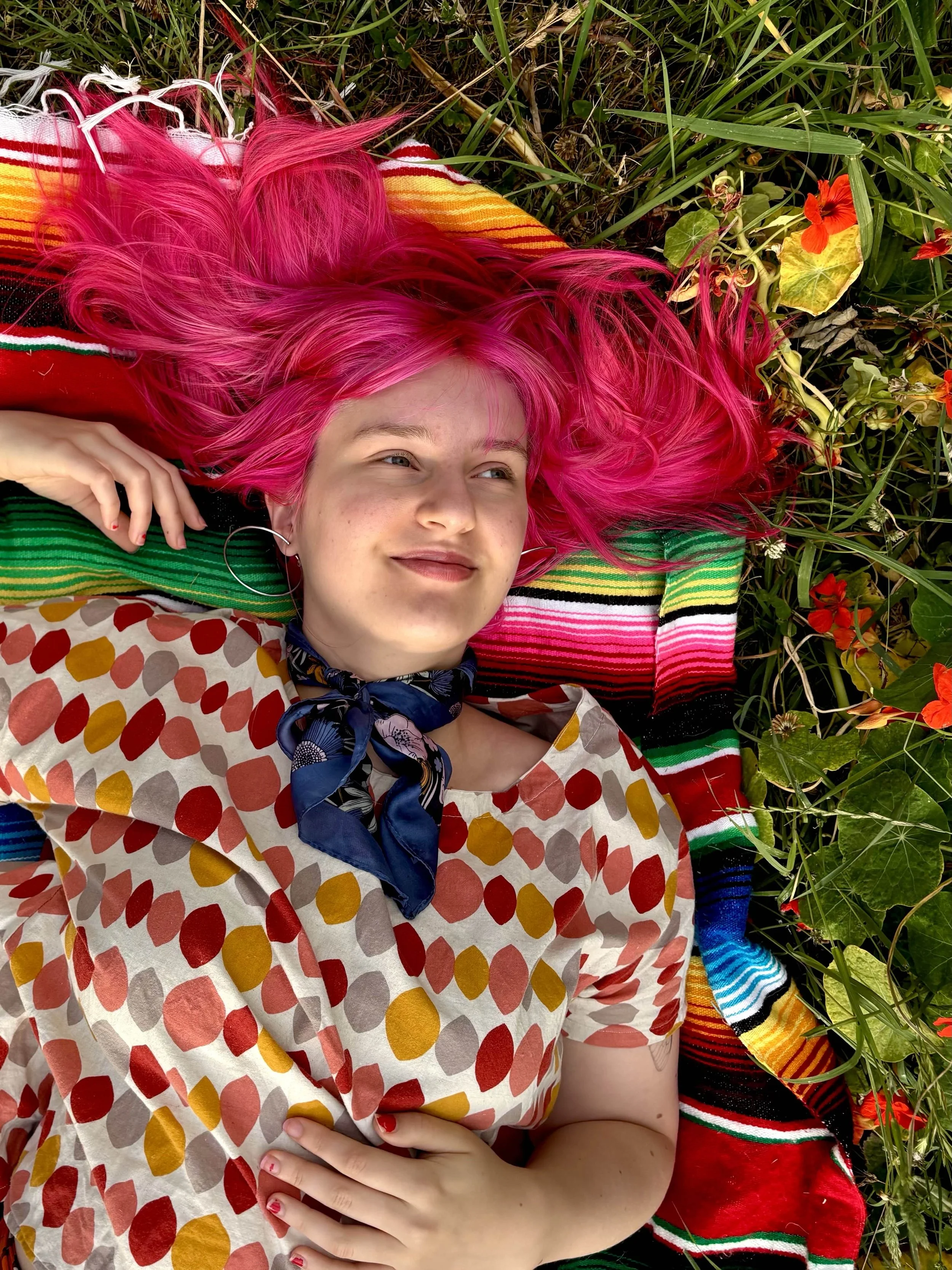 Audrey with bright pink hair lying on a striped blanket among green grass and orange flowers.