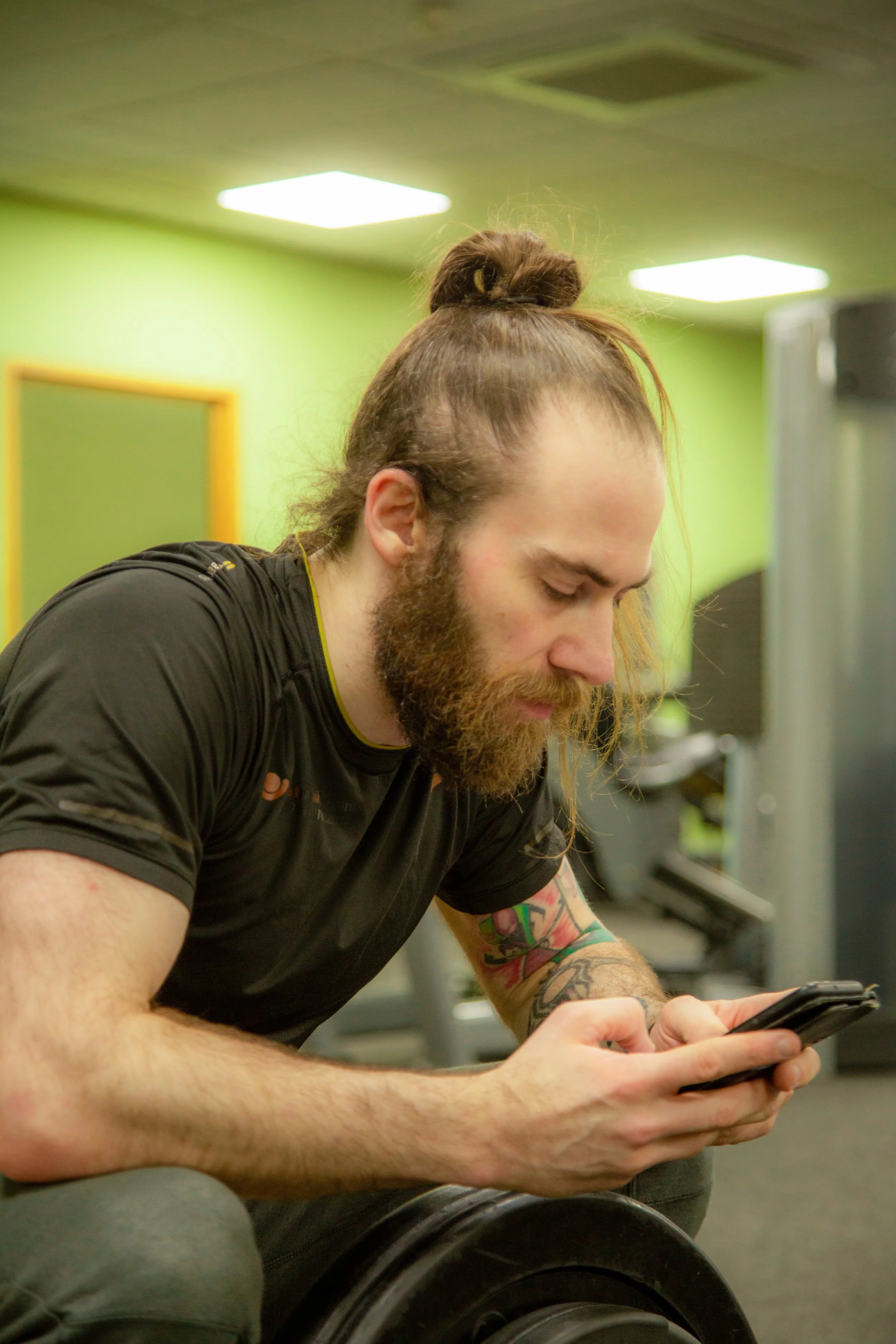 Photo of the practitioner: a man with long hair tied in a bun and a beard, wearing a black athletic shirt, sits in a gym. He has colourful tattoos on his arm, and the background features green walls and ceiling lights.