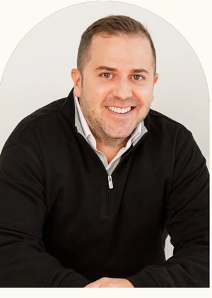 A smiling man with short hair, wearing a black sweater over a collared shirt, seated with his arms on a table against a white background.