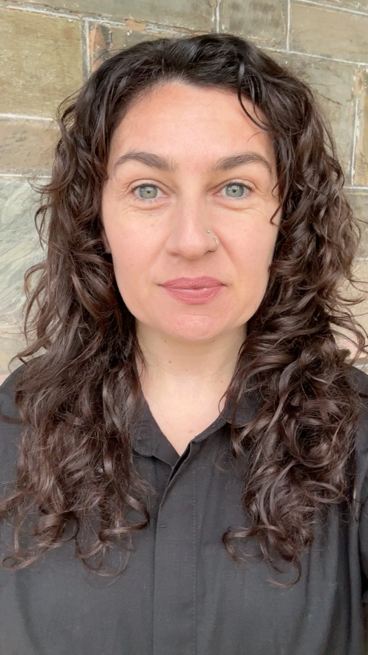 A woman with curly brown hair and light skin, wearing a black shirt, standing against a brick wall.