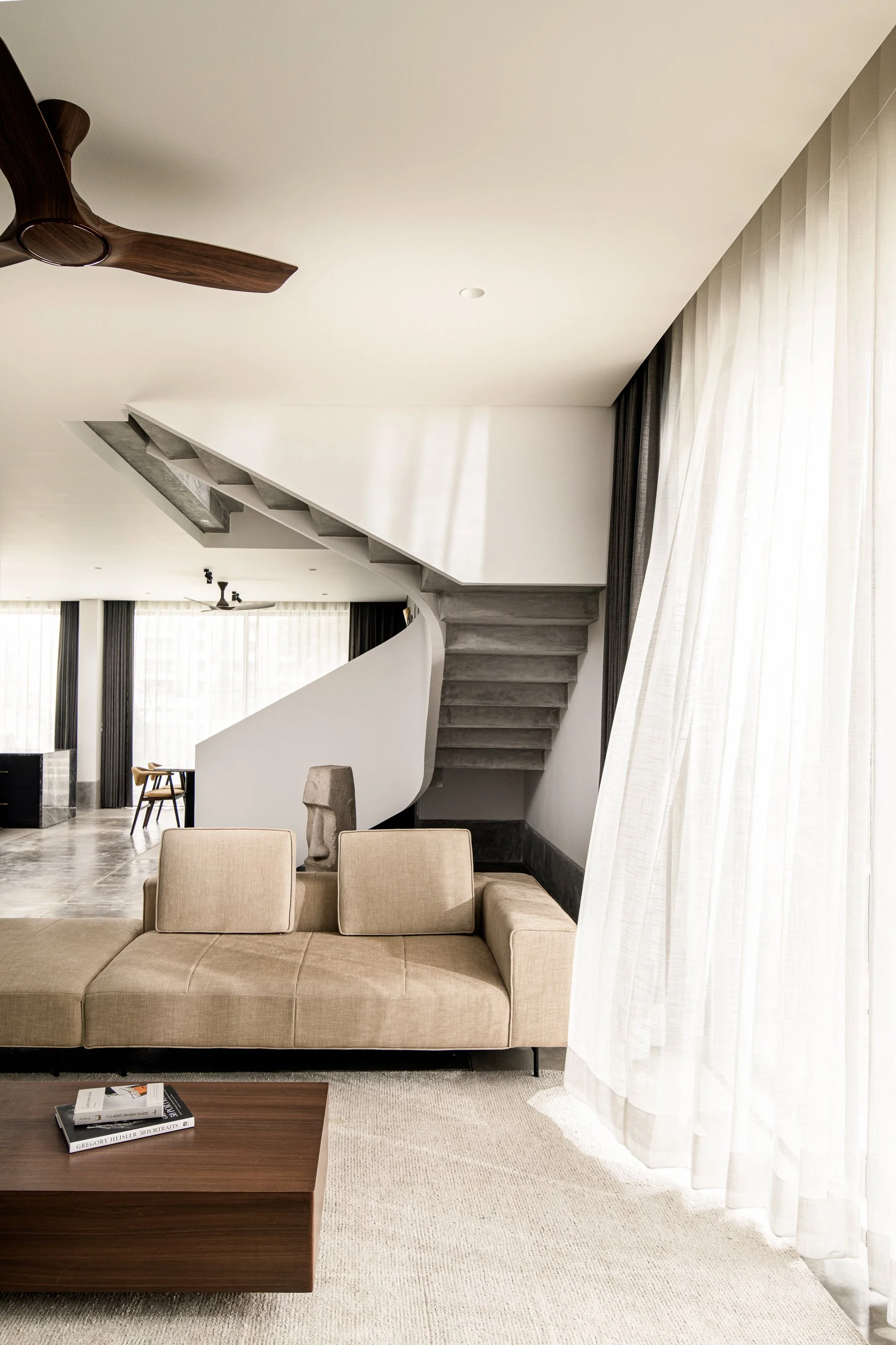 Modern living room with beige sofa, wooden coffee table, abstract sculpture, staircase, and large windows with white curtains.