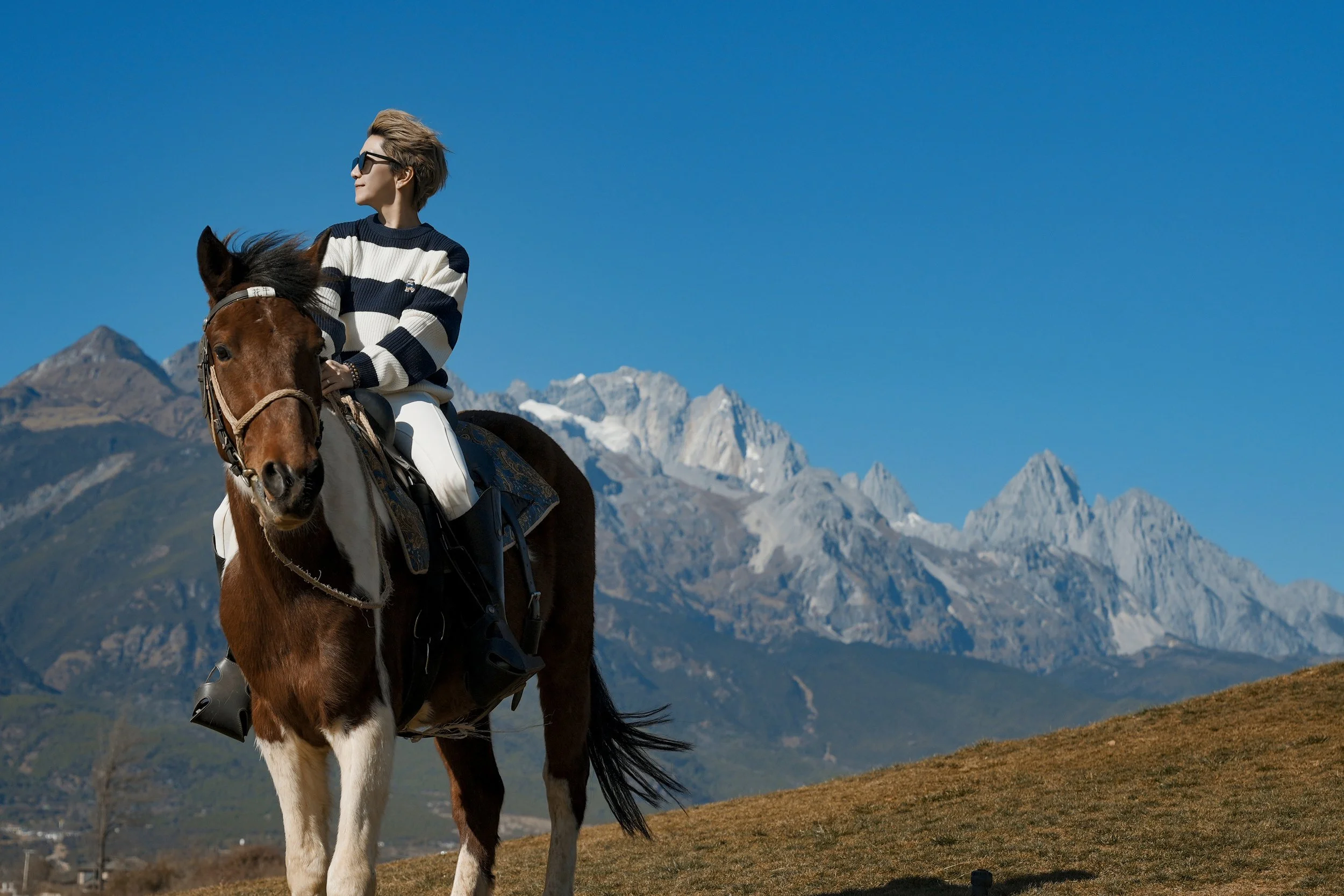 Person riding a brown and white horse in a mountainous landscape with clear blue sky.