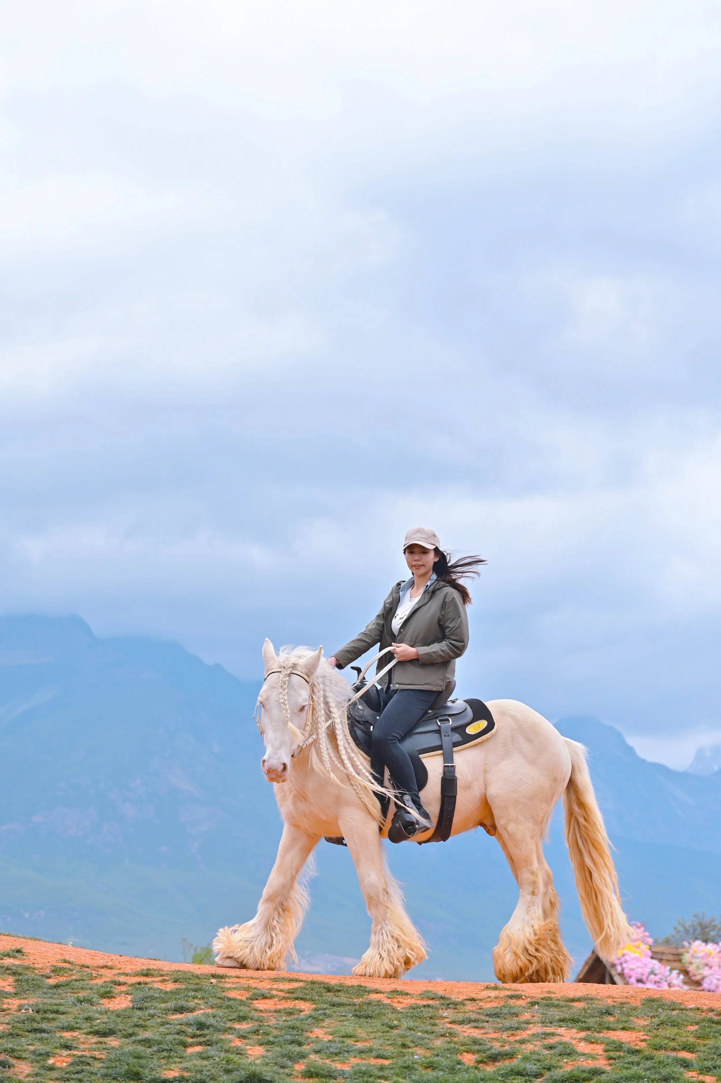 A woman riding a white horse with a mountain landscape and cloudy sky in the background. The horse has long, flowing hair and the woman is wearing casual outdoor clothing, including a gray cap and jacket.