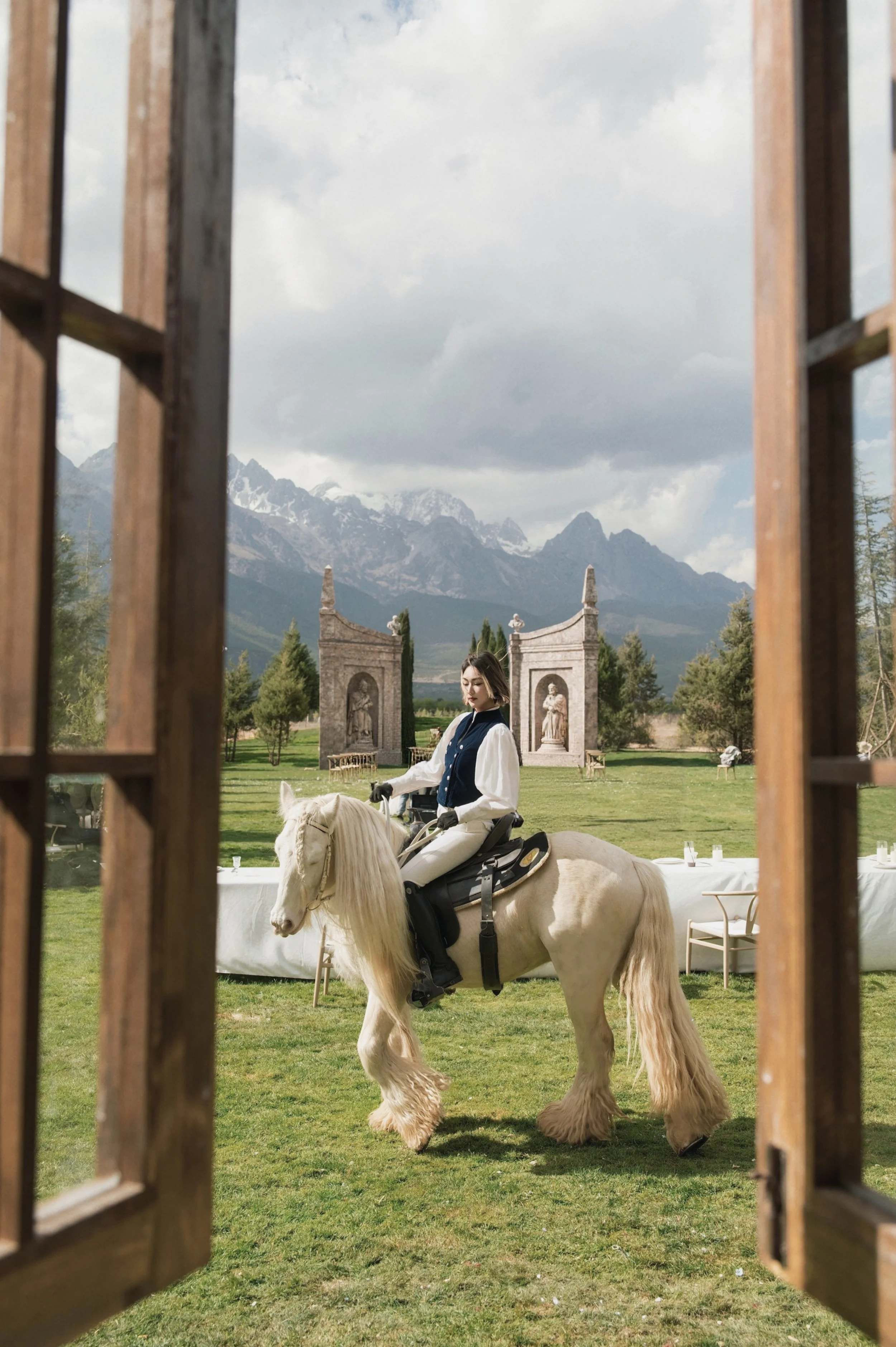 A woman riding a white horse, seen through a wooden window frame, with mountains and trees in the background.