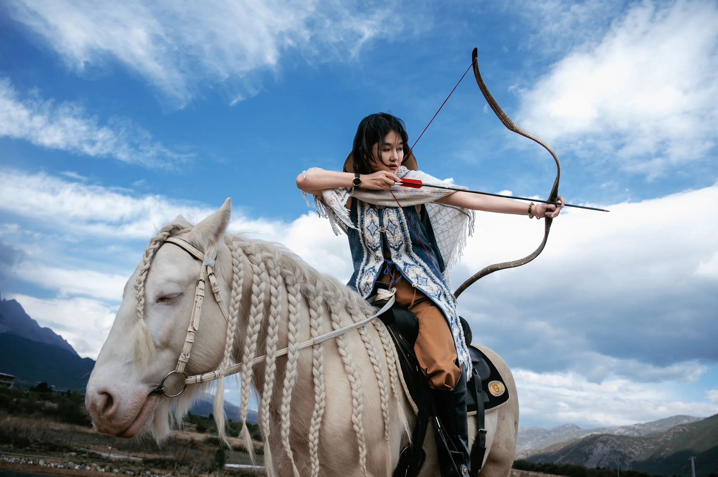 A woman riding a white horse with braided mane, aiming a bow and arrow towards the sky in a scenic outdoor landscape with mountains and blue sky with clouds.