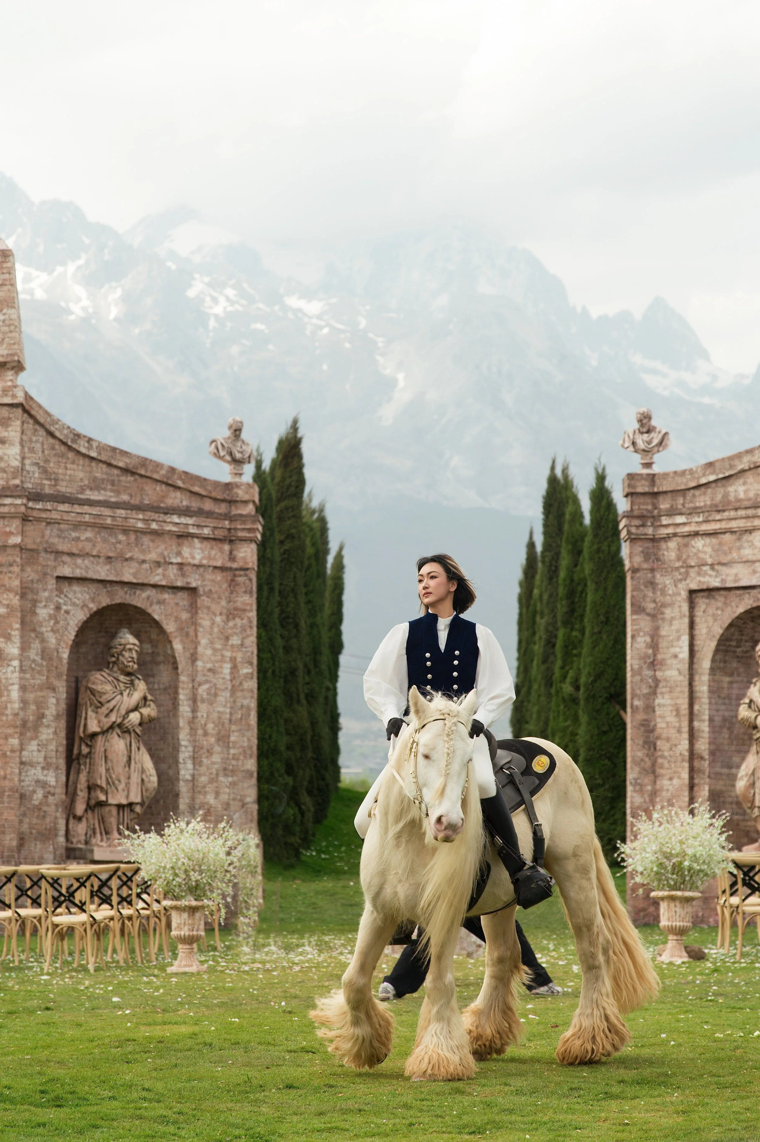 A woman on a white horse in front of stone archways with statues, green trees, and mountains in the background.