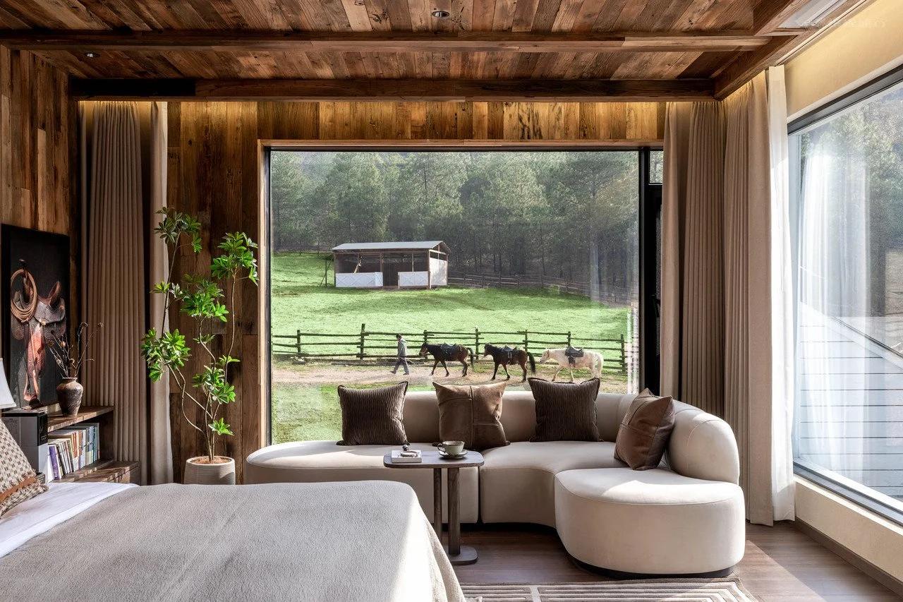 Living room with large window view of green backyard, with horses and a person walking, wooden walls, and a white curved sofa with pillows.