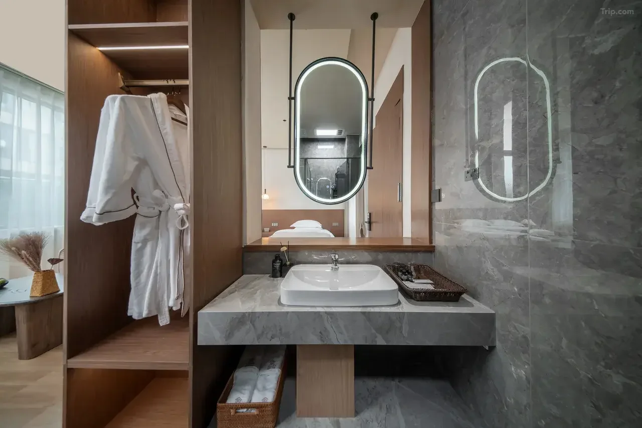 Modern bathroom with a marble sink, an illuminated oval mirror, and a wooden wardrobe with a white bathrobe hanging inside. There is a window with sheer curtains on the left and a bedroom with a bed visible through the mirror reflection.