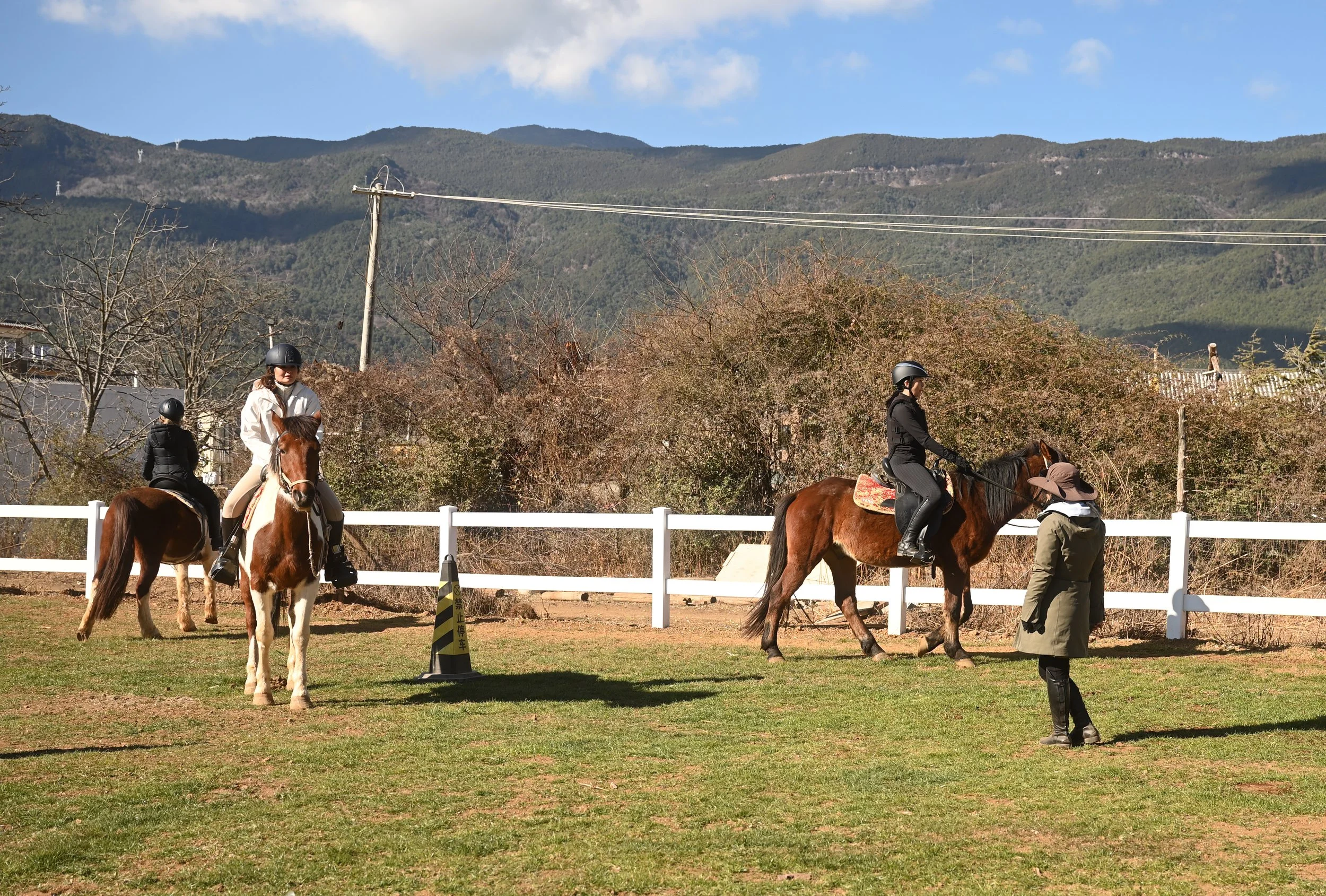 Several horseback riders wearing helmets riding on a field with white fencing, with trees and mountains in the background under a partly cloudy sky.