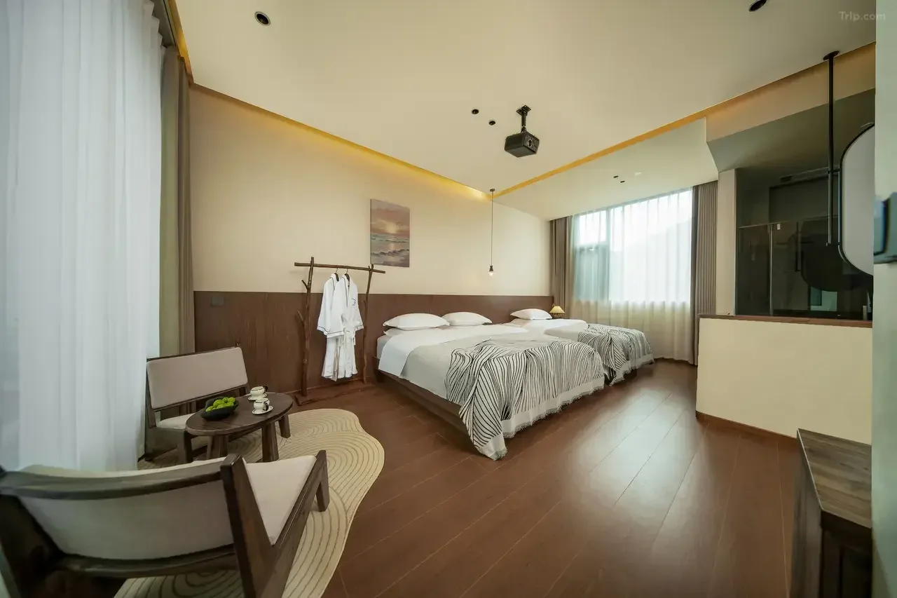 Hotel room with three beds, a sitting area with two chairs and a small table, and a window with curtains. A bathrobe hangs on a wooden coat rack.