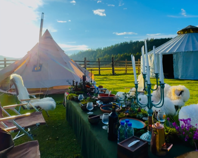 Outdoor scene with a large table set for a meal, decorated with a candelabrum, bottles, and tableware, in front of two tents, one beige and the other white, on a grassy field with a wooden fence and green hills in the background under a sunny sky.