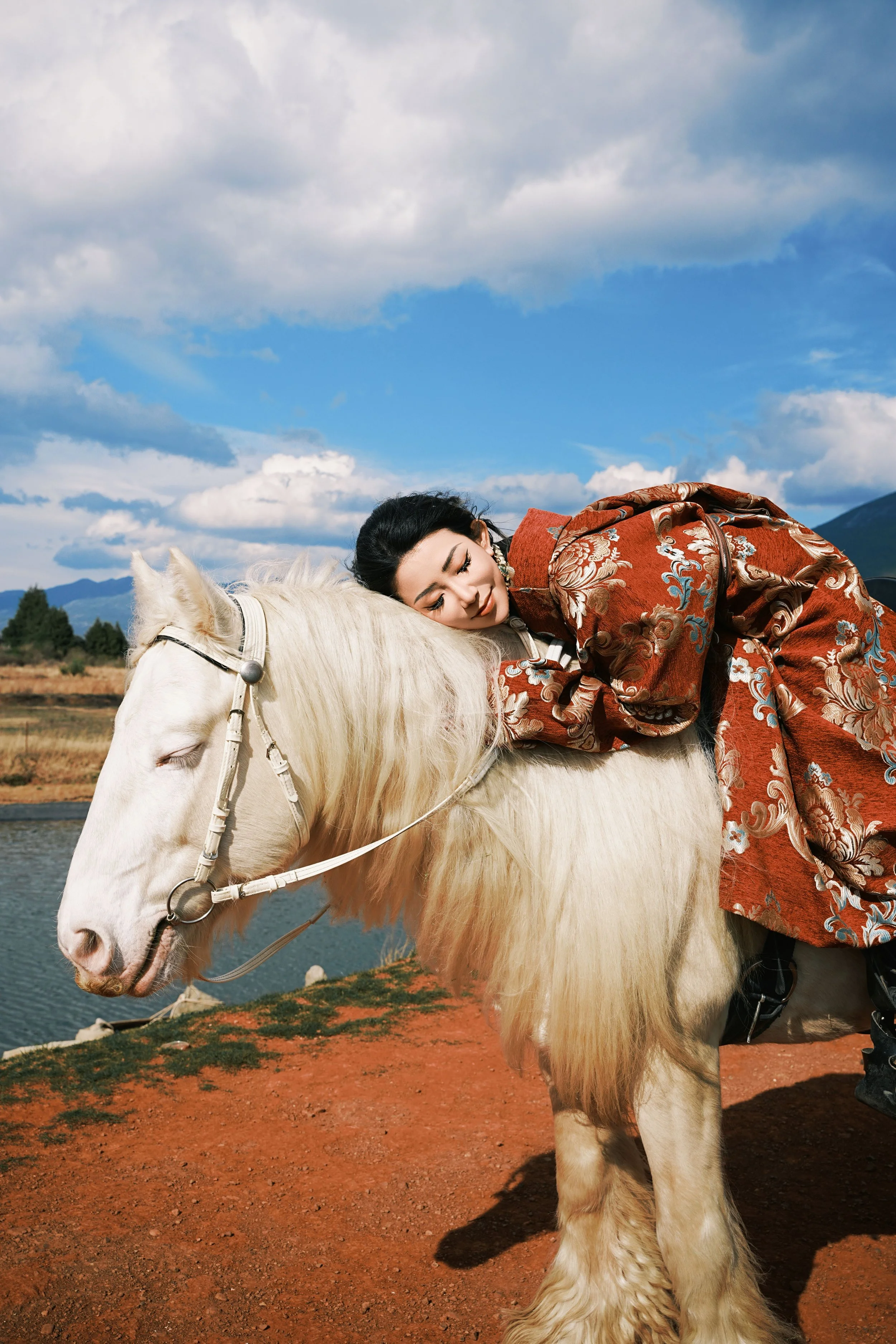 A woman in traditional attire resting on a white horse in an outdoor landscape with a river, trees, mountains, and partly cloudy sky.