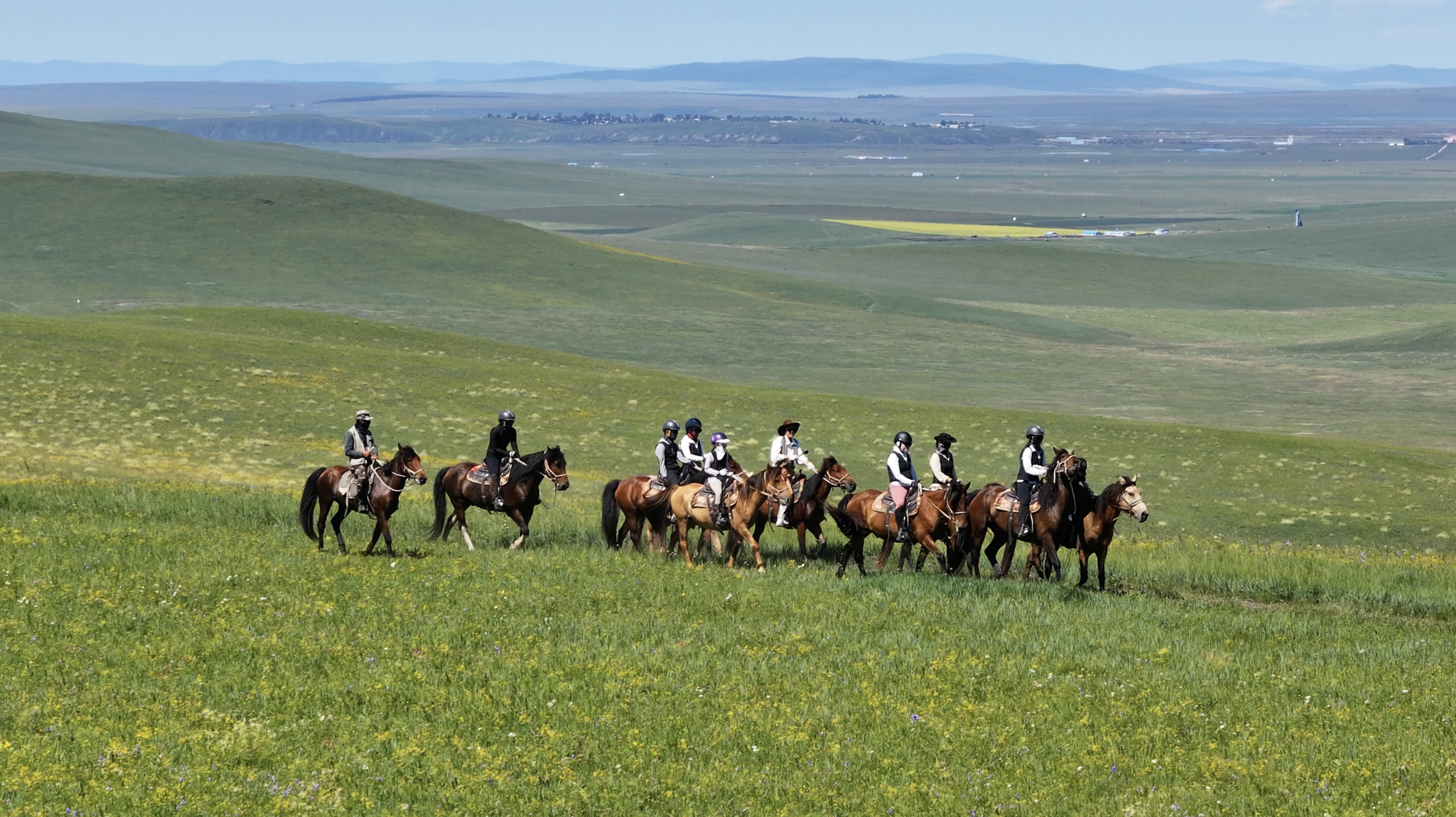 A group of ten people riding horses across a green, grassy field with rolling hills and distant mountains in the background.