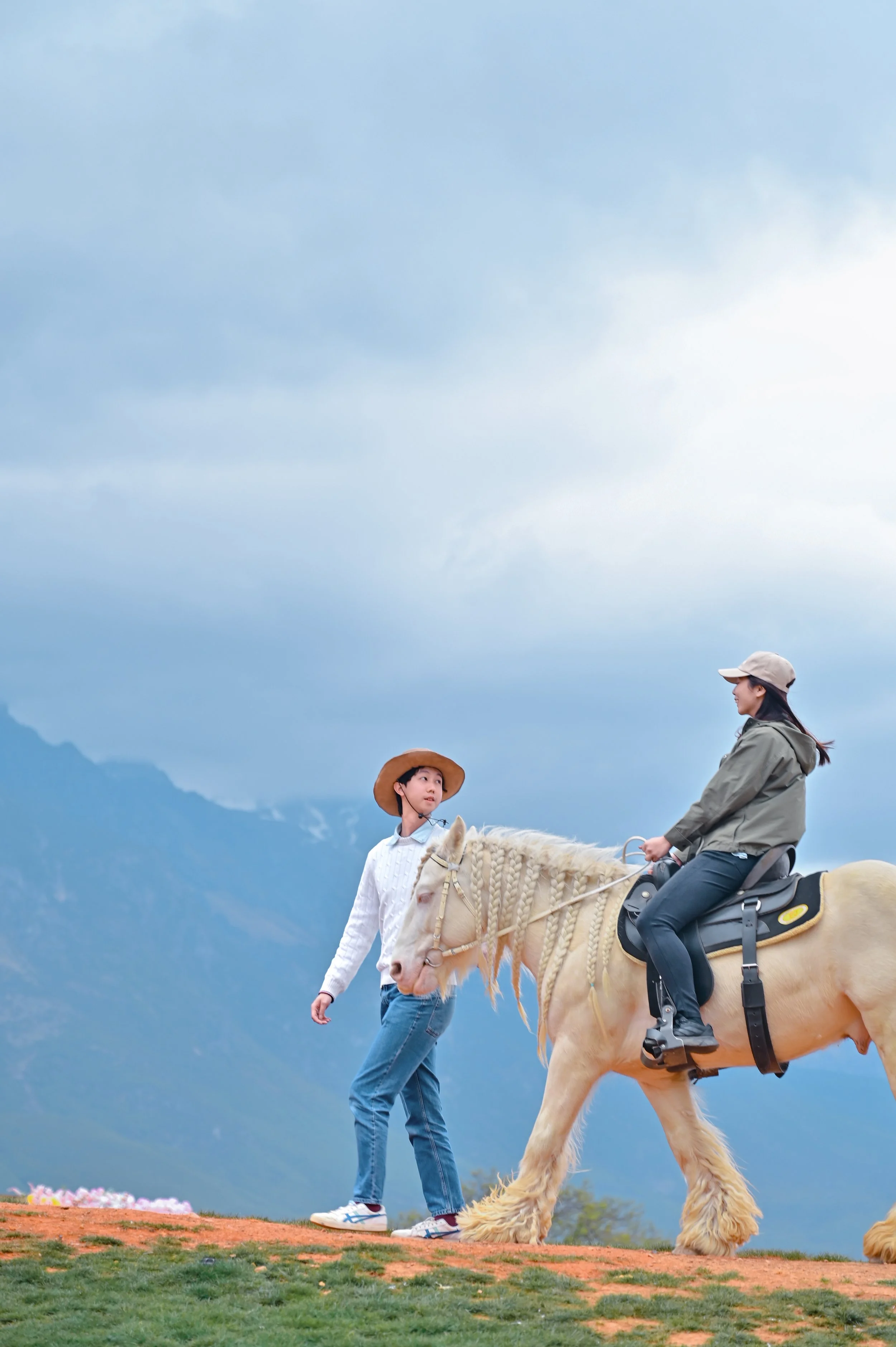 A woman riding a large beige dog with long fur, wearing a saddle, on a grassy hill. A young boy with a wide-brimmed hat and a woman in a beige cap stand nearby. The background features a mountain range under a cloudy sky.