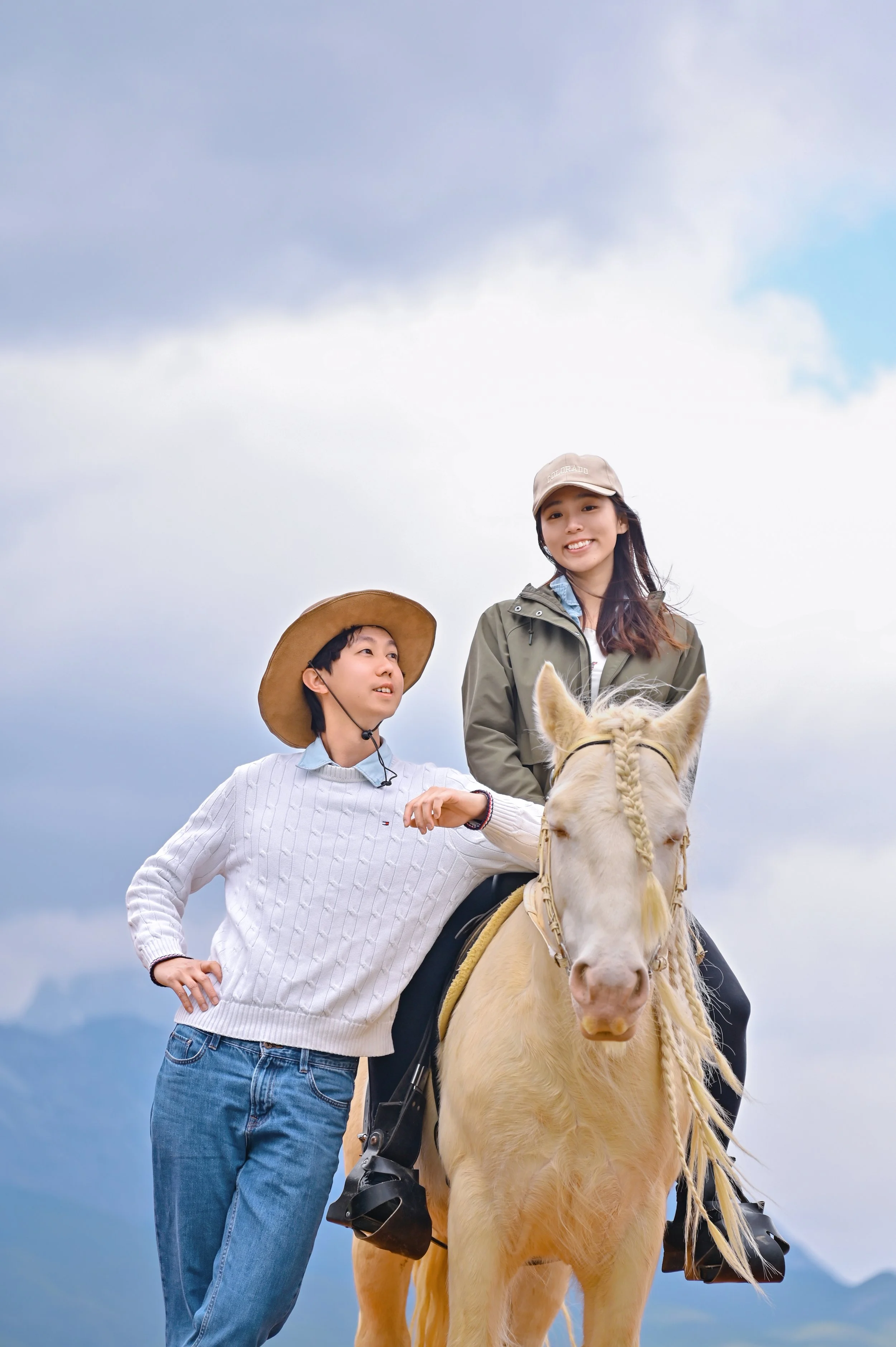Two young women, one in a white sweater and the other in a green jacket, riding a light-colored horse with a blonde mane. The woman in the jacket is smiling, while the woman in the sweater is looking at her. They are outdoors under a cloudy sky, with