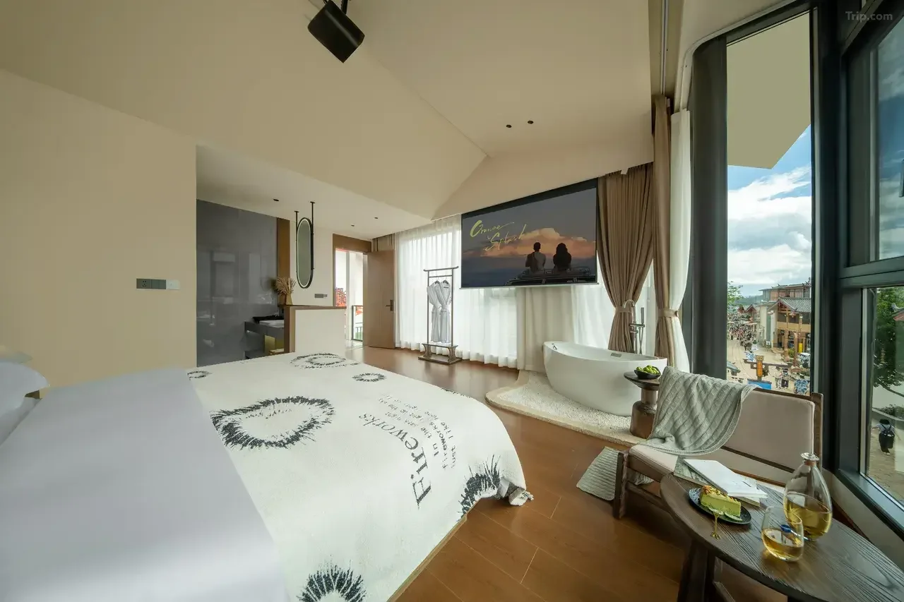 A modern hotel room with a large bed, a flat-screen TV, a freestanding bathtub near floor-to-ceiling windows, and a small sitting area with a table holding a bottle of wine and glasses, overlooking a street outside.