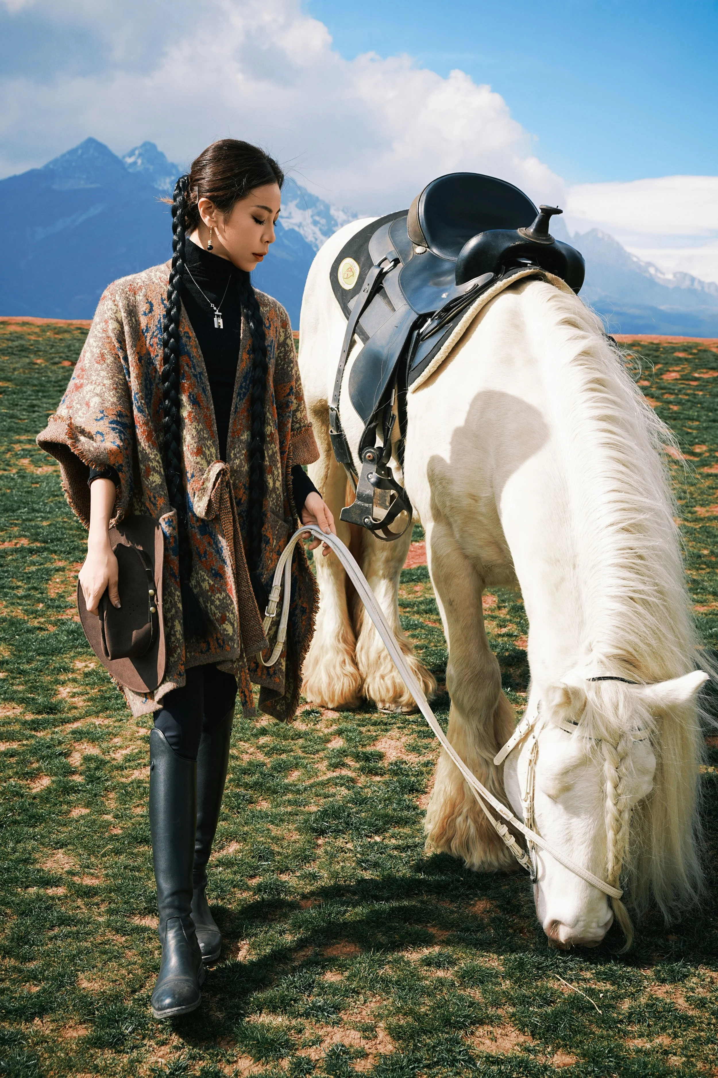 A woman with dark hair in braids, wearing a patterned coat and black boots, standing next to a white horse with a black saddle in a grassy field with mountains and blue skies in the background.