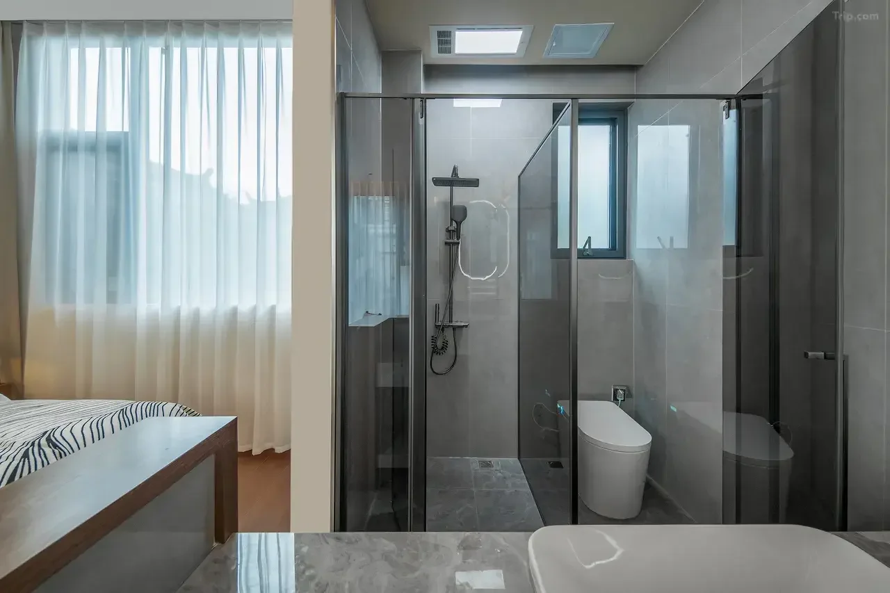 Modern bathroom with glass shower enclosure, toilet, small window, and marble-like wall tiles, visible from a nearby bedroom with curtain and bed.