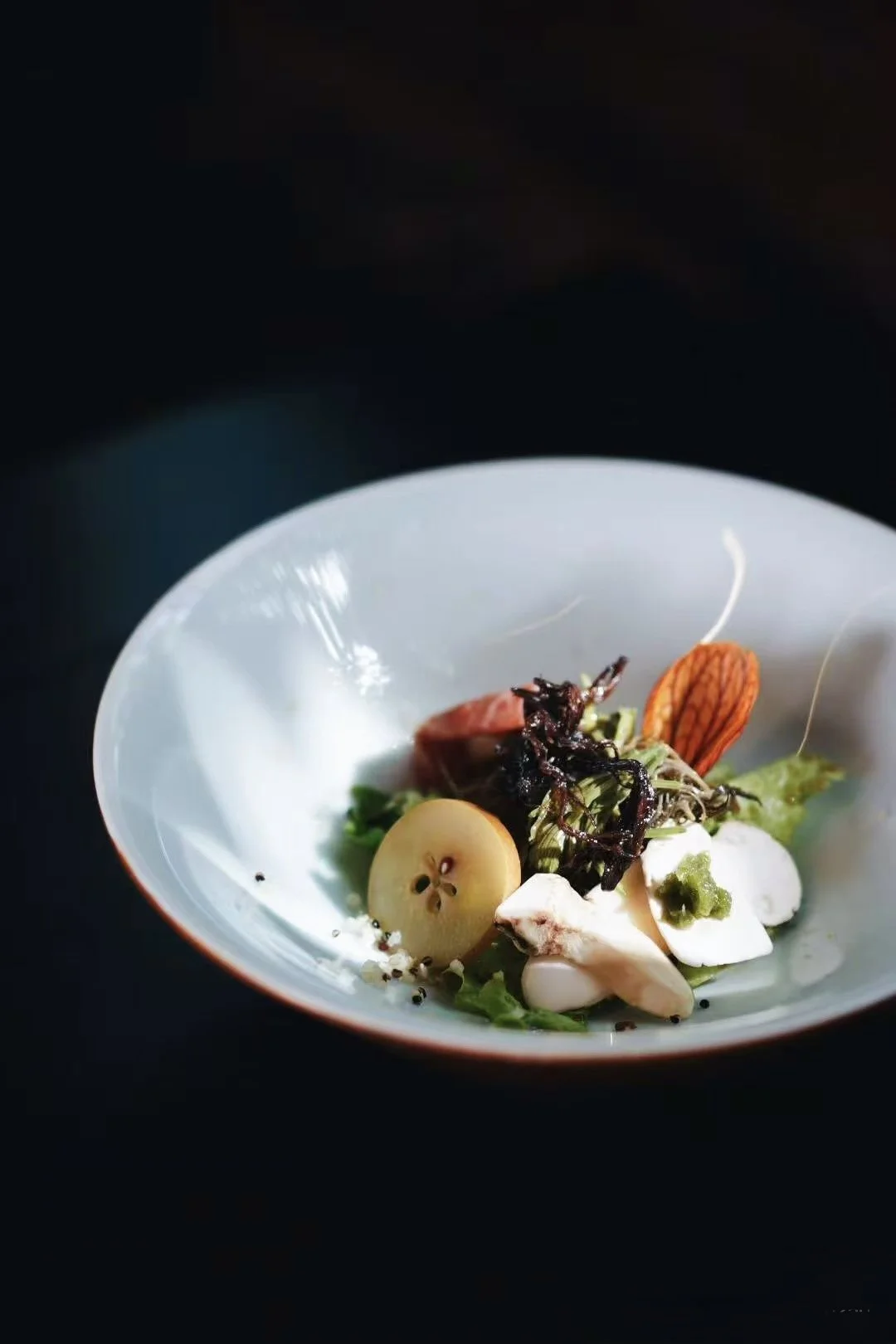 Elegant plated dish with various vegetables, greens, and garnishes on a white plate, against a dark background.