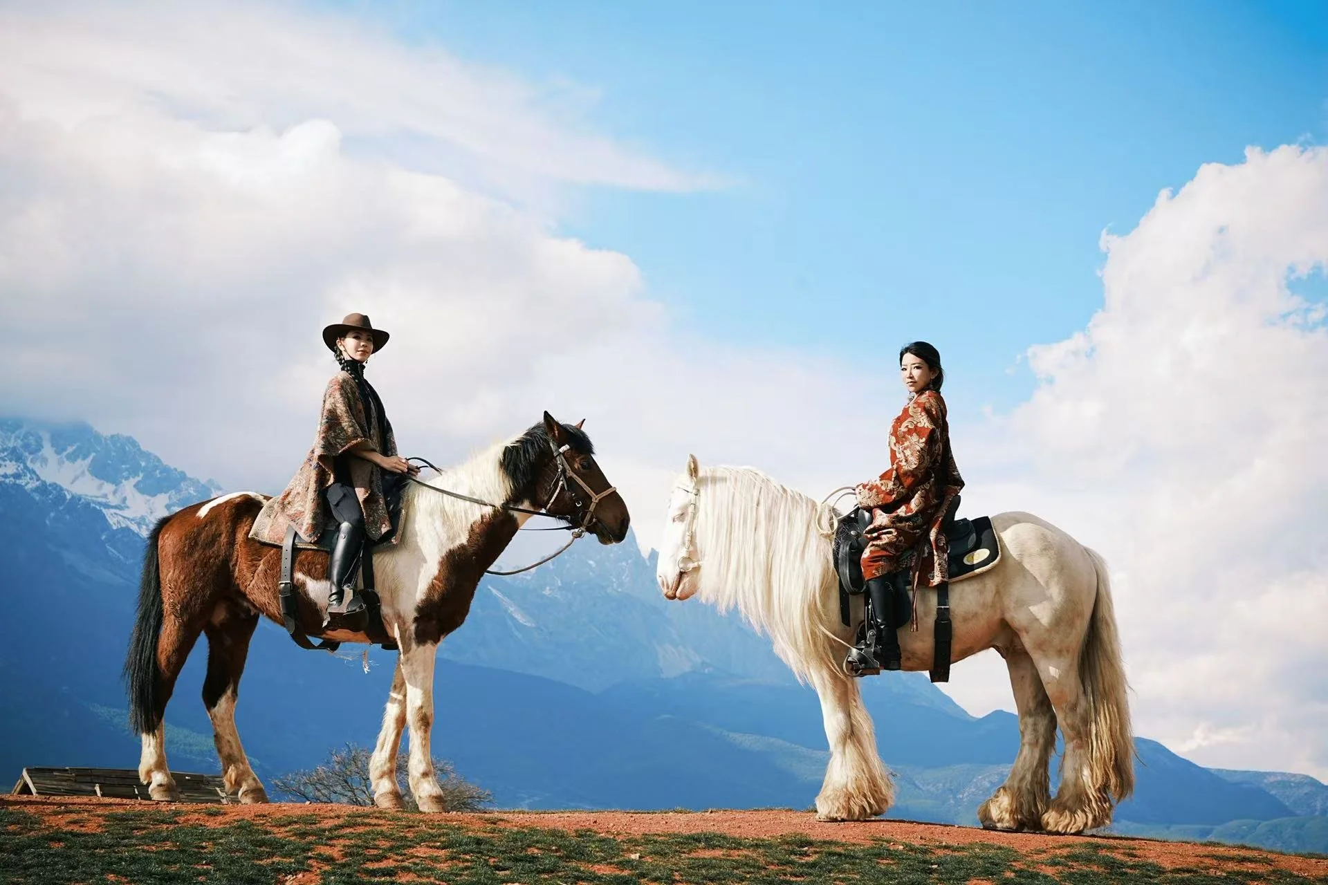Two women riding horses outdoors with mountains and cloudy sky in the background.