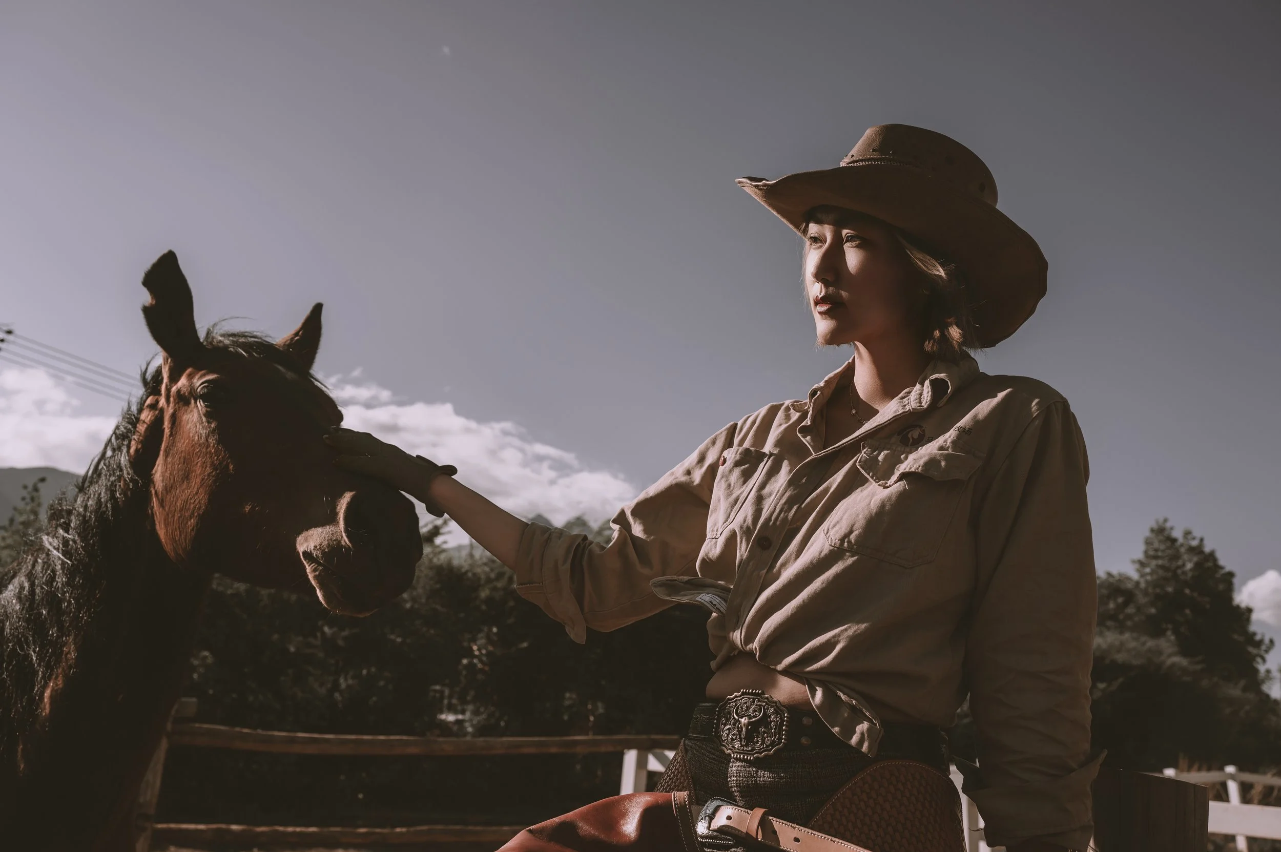 A woman wearing a cowboy hat and western attire stands next to a brown horse in a rural outdoor setting during daytime.