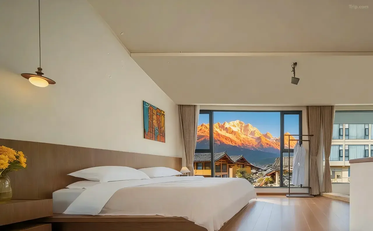 Hotel room with large window showing mountain landscape at sunrise, king-sized bed, artwork on wall, flower vase, and clothing rack.