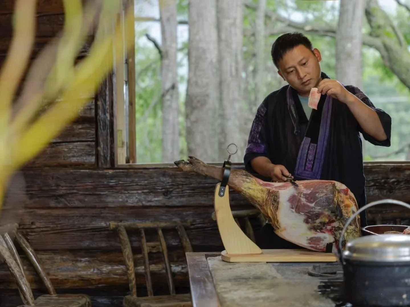 A person uses a knife to cut a large piece of cured ham or prosciutto on a wooden stand with a scenic outdoor background visible through a window.