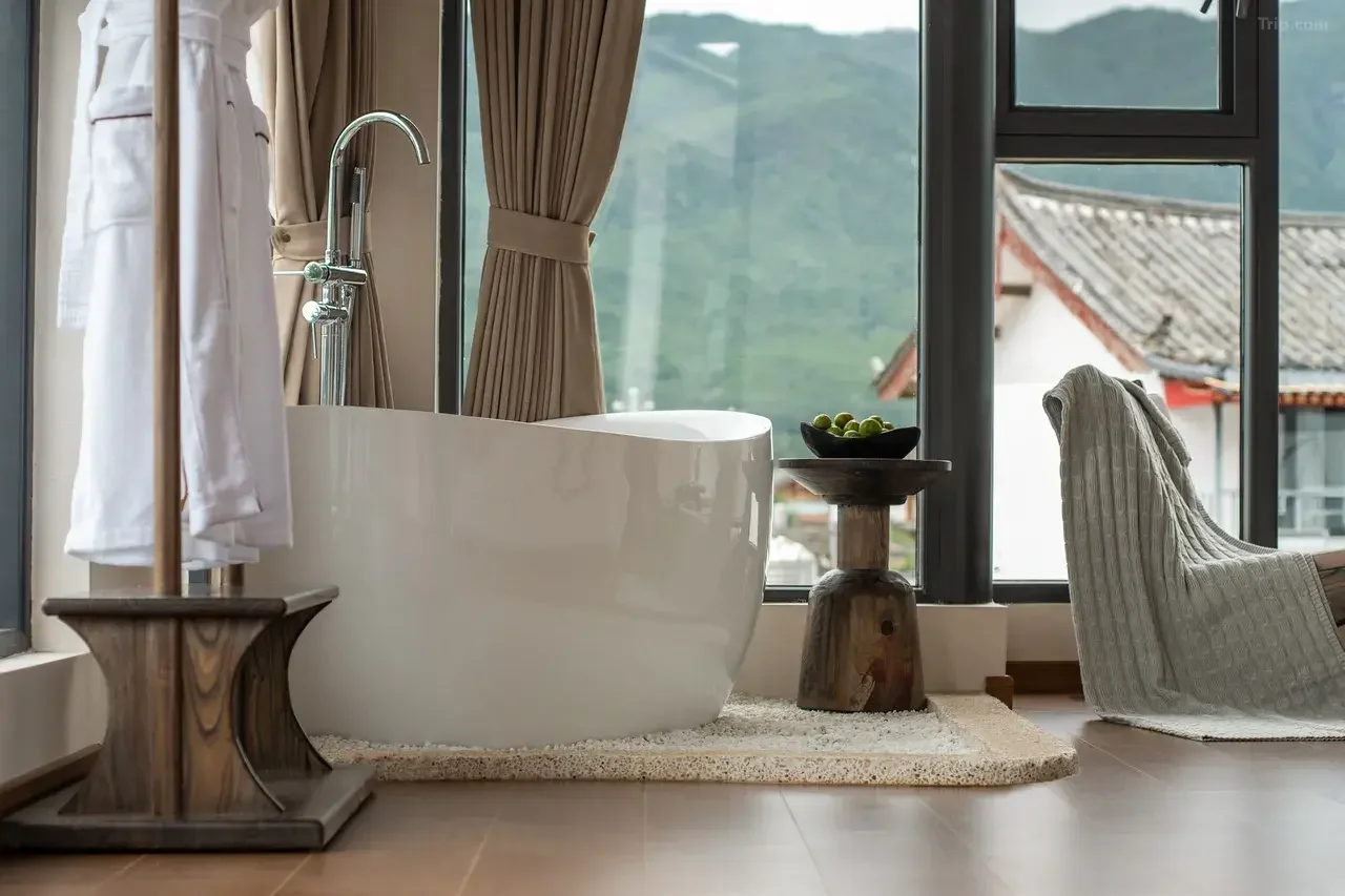 A modern bathroom with a white bathtub near large windows with beige curtains, a wooden stool with a bowl of green fruit, a cushioned chair covered with a grey blanket, and views of traditional rooftops outside.