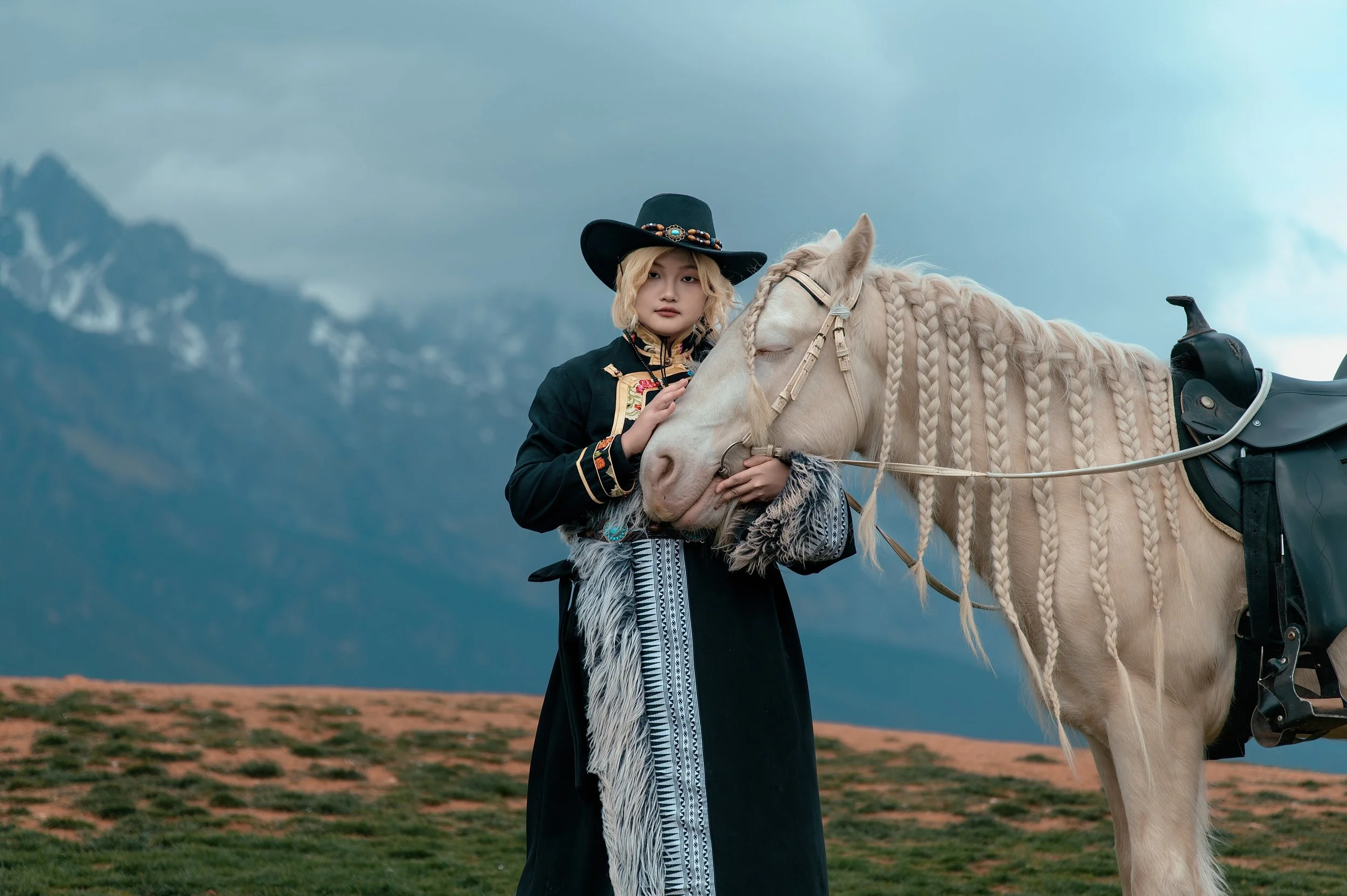 A young woman with blonde hair wearing traditional clothing and a black hat standing next to a white horse with braided mane in an outdoor landscape with mountains in the background during cloudy weather.