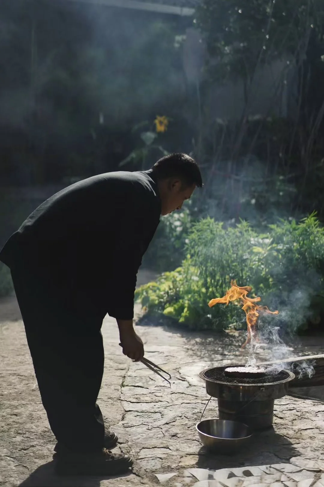 A man dressed in black clothes is tending a small outdoor fire on a metal grill, with flames and smoke rising, surrounded by greenery on a stone patio.