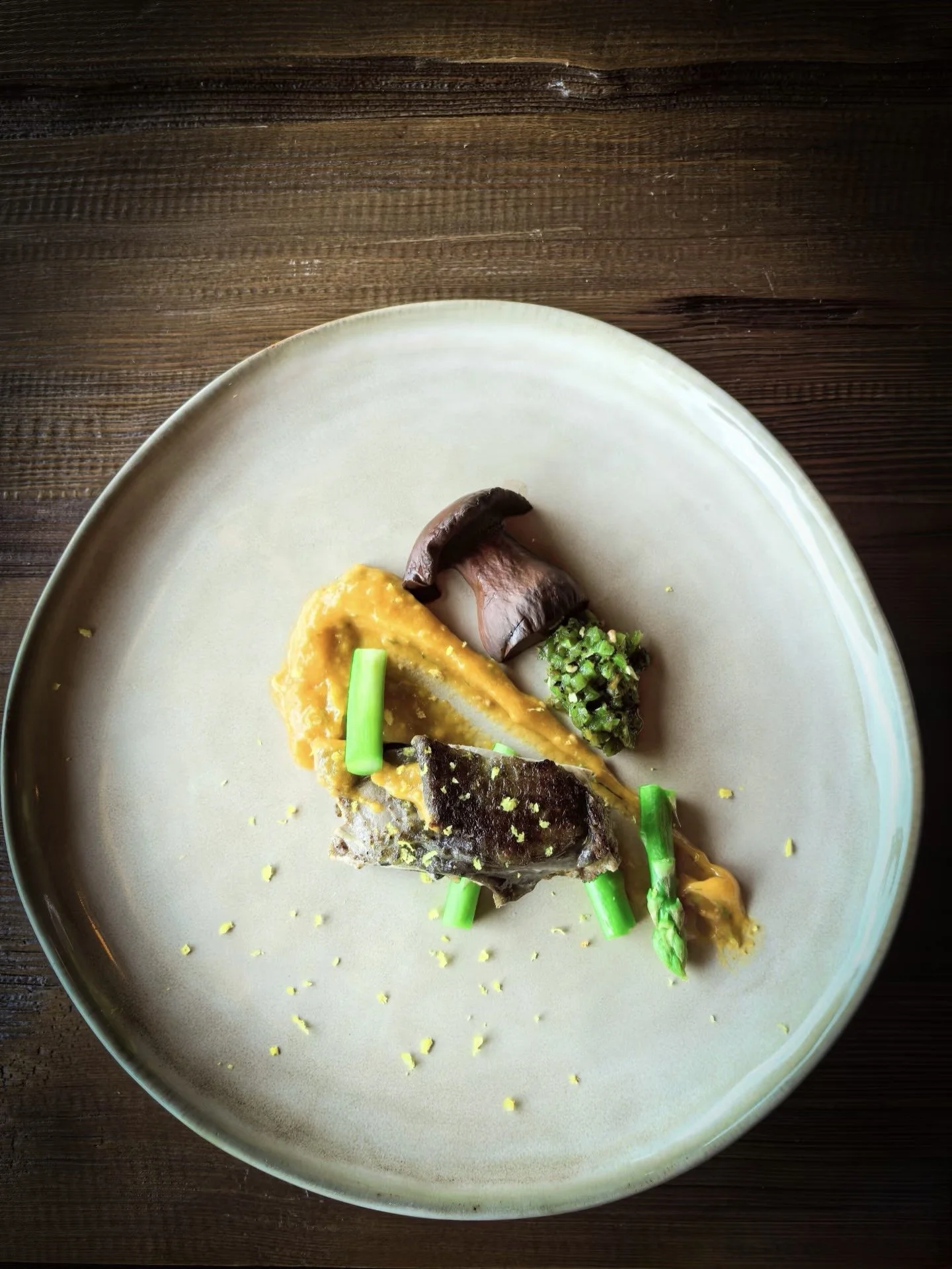 A plate with a serving of grilled fish topped with a yellow sauce, garnished with green vegetables and small pieces of black seaweed, on a wooden table.