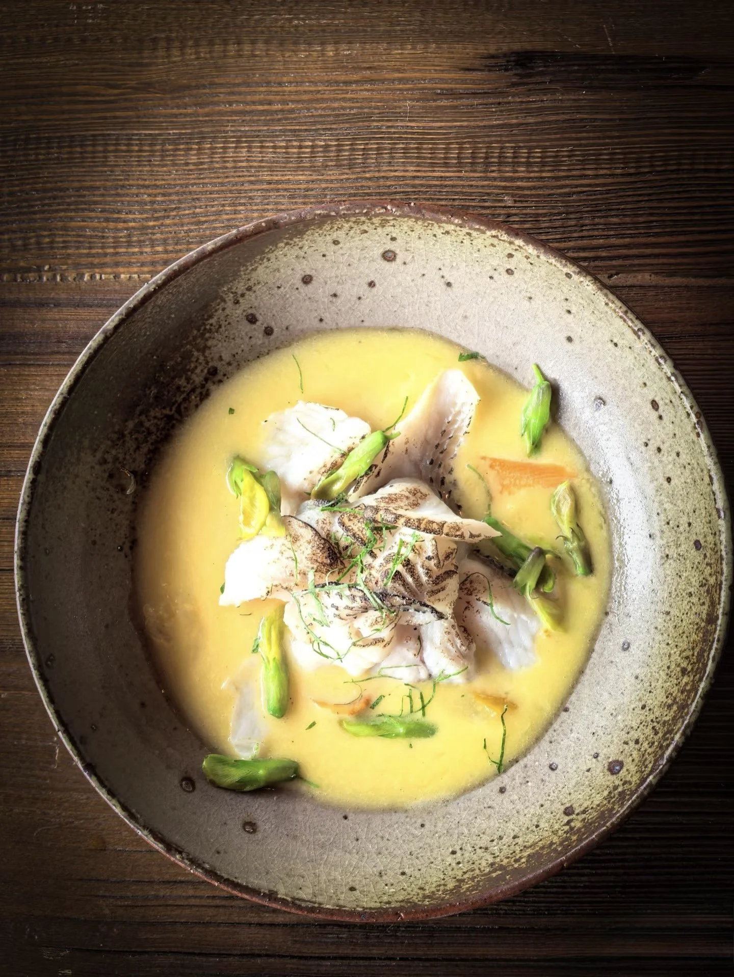 A bowl of yellow soup with pieces of fish, green vegetables, and garnishes on a wooden table.