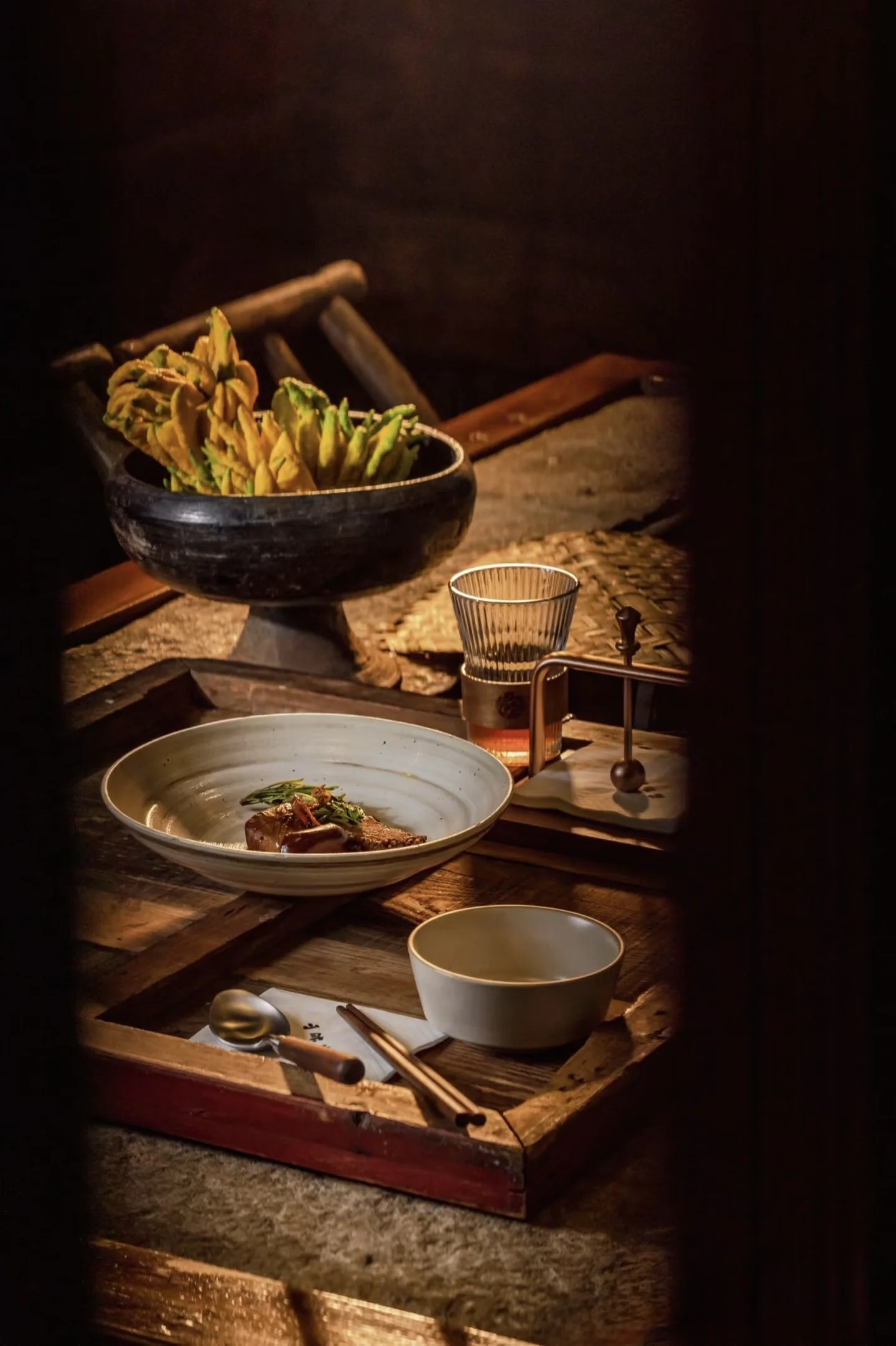 A rustic wooden dining table set with Asian-style dishes, including a bowl of vegetables, a plate with meat and herbs, a glass of beverage, and traditional chopsticks and utensils in warm lighting.
