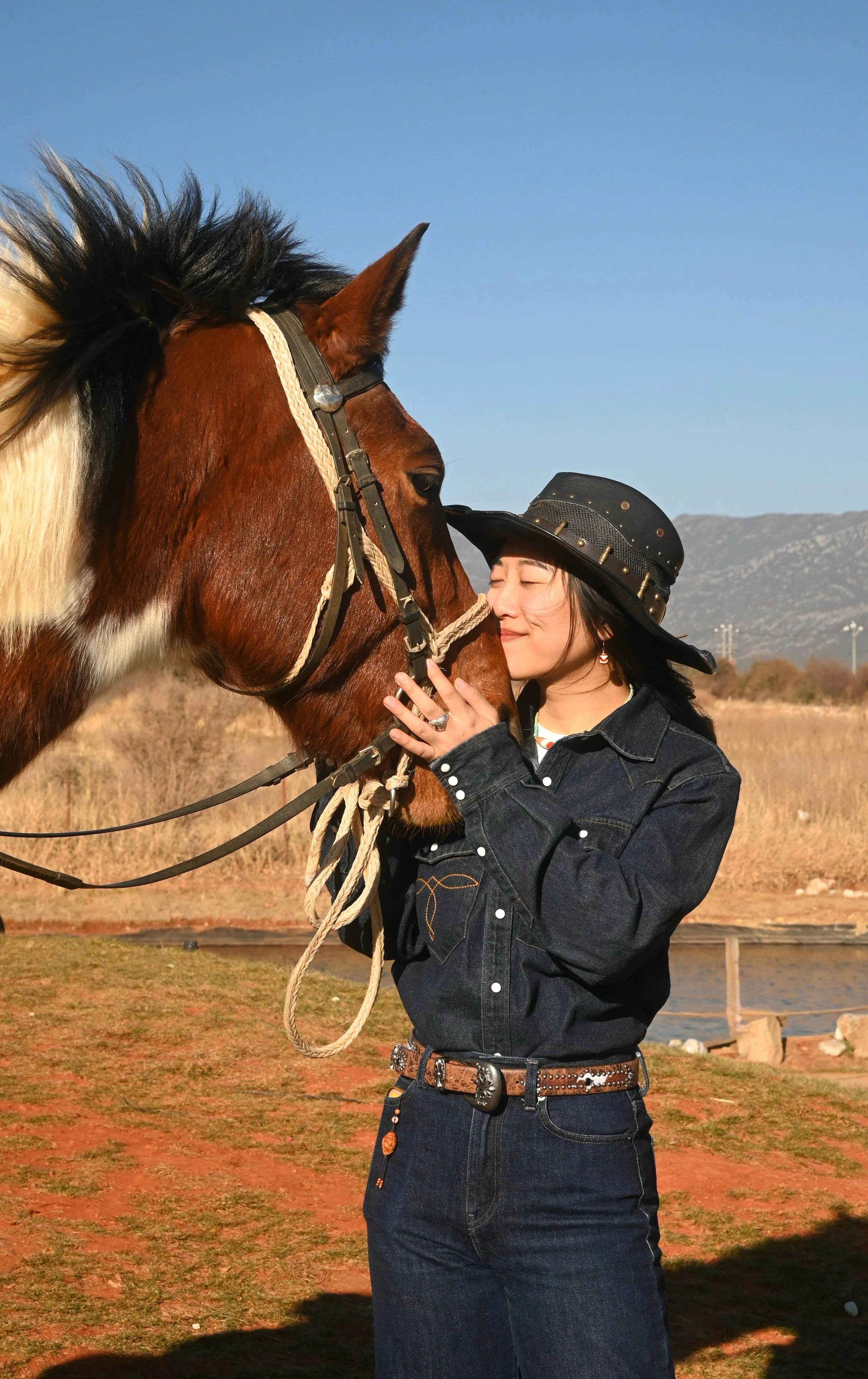 A young woman with long dark hair in a cowboy hat smiling with eyes closed, embracing a brown and white horse outdoors during daytime.