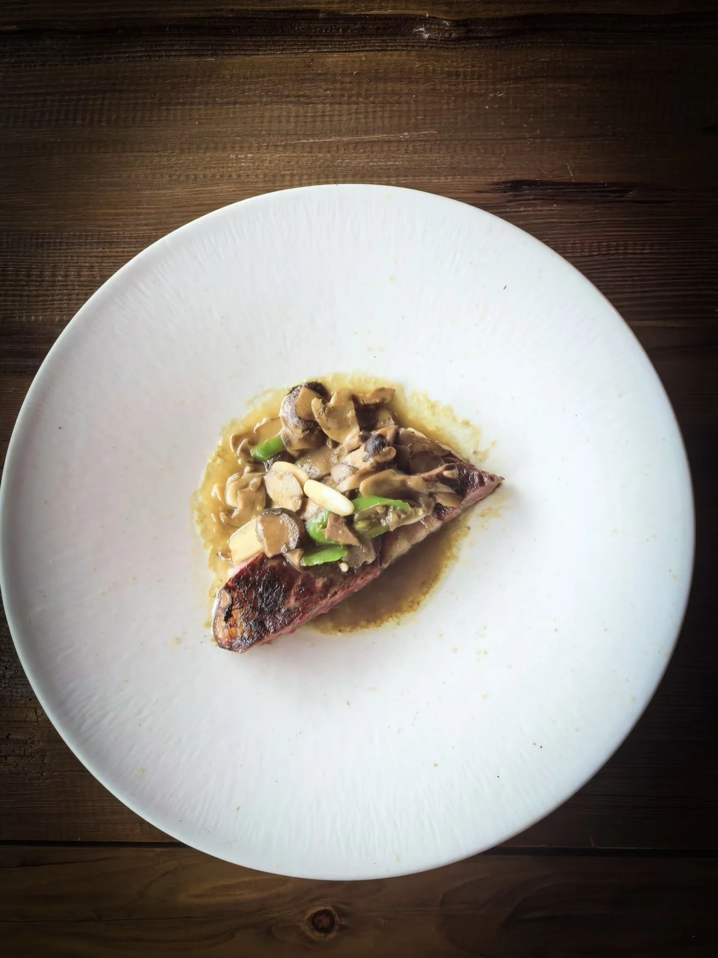 A piece of cooked fish topped with chopped mushrooms and green herbs on a white plate on a wooden table.
