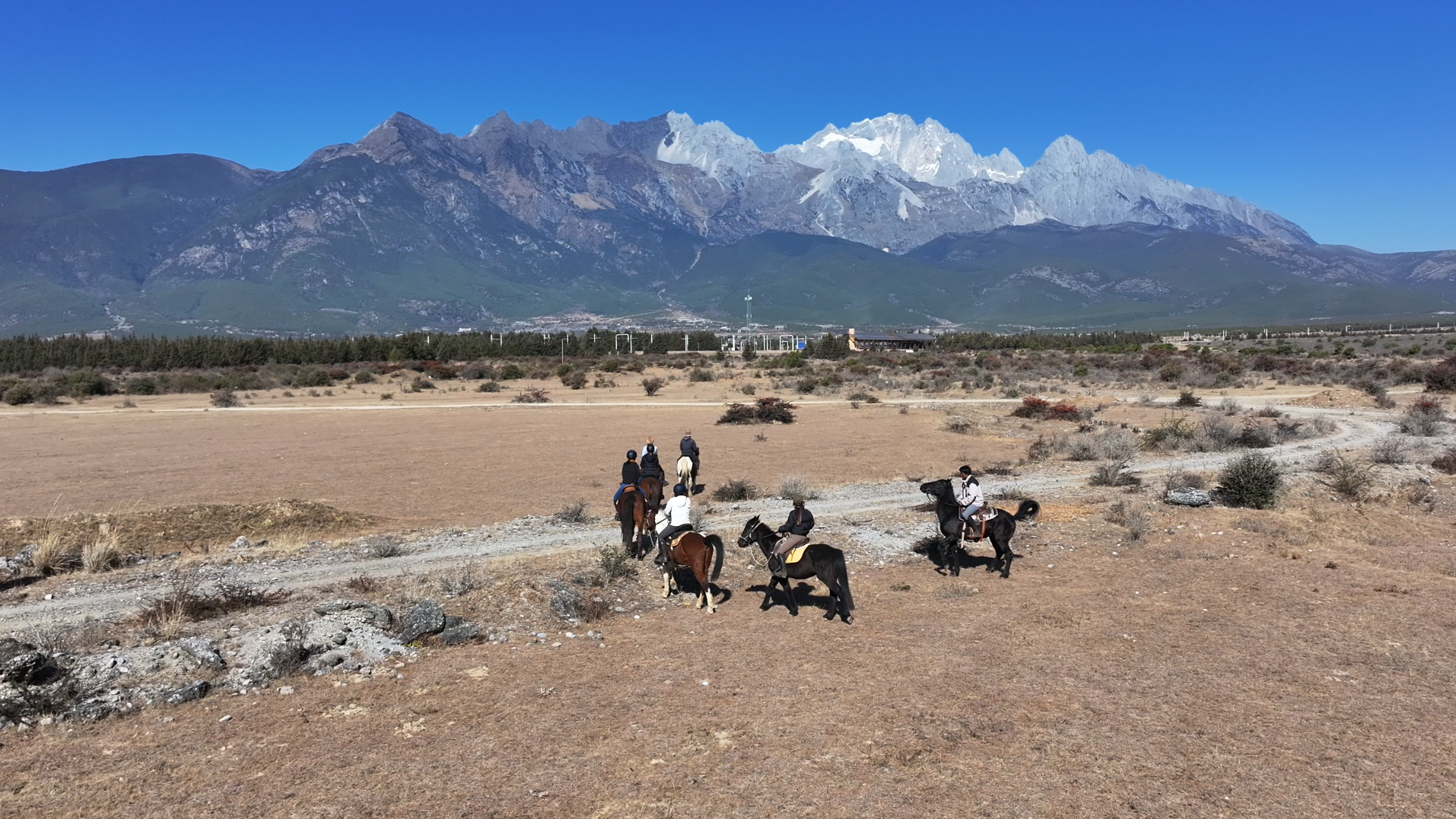Group of people horseback riding on a dirt trail in a barren landscape with mountains in the background and a clear blue sky.