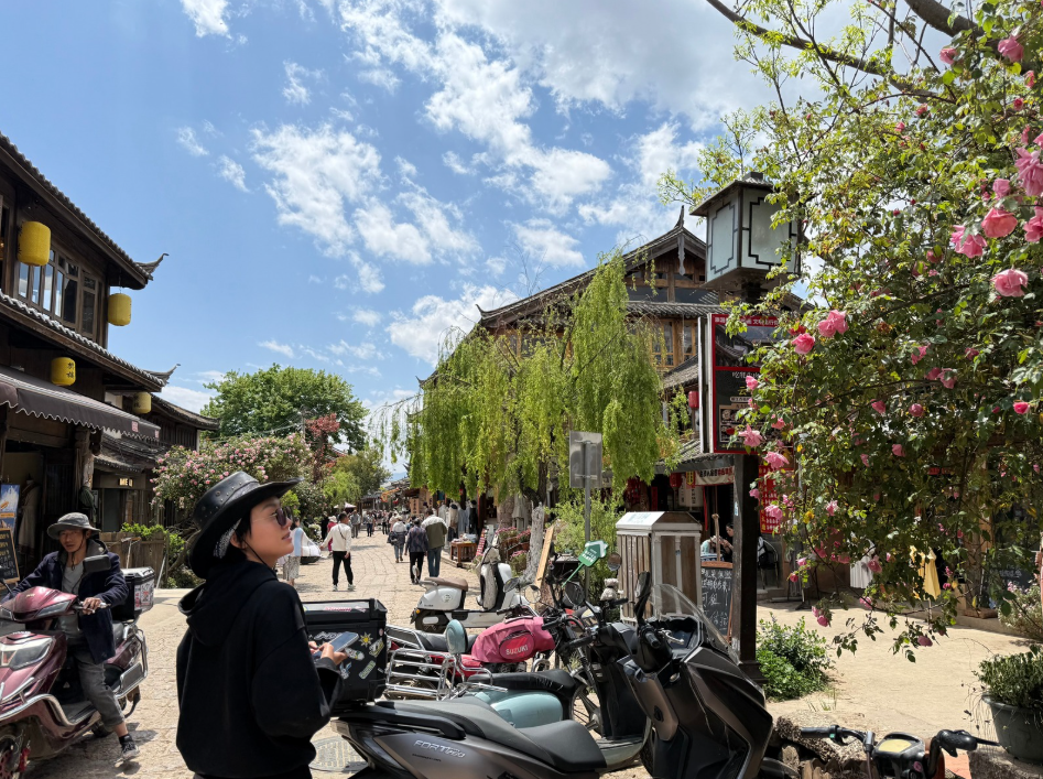 People walking and riding scooters on a bustling street with traditional Asian buildings, colorful flowers, and a partly cloudy sky.