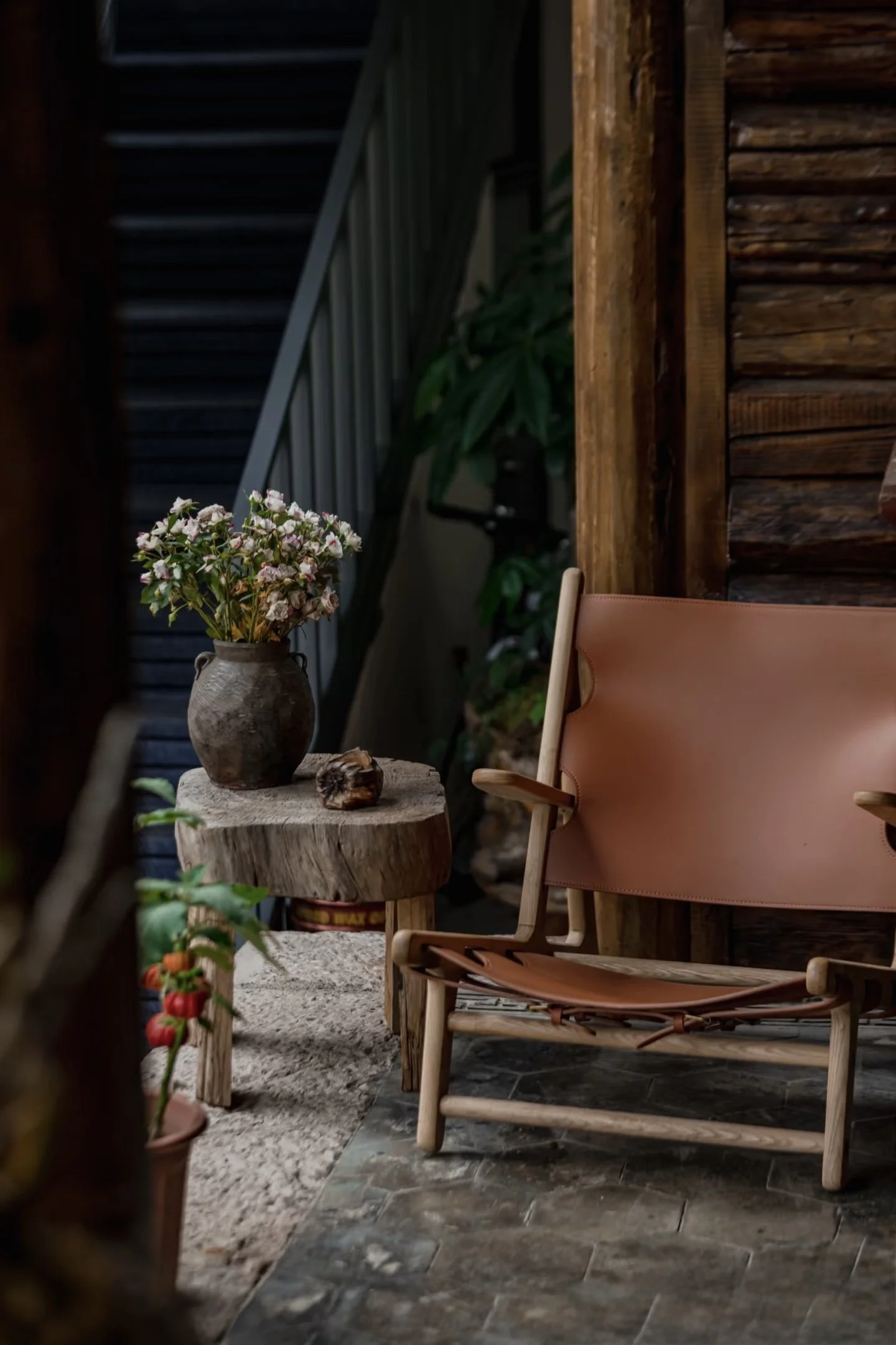 Rustic indoor or porch setting with a brown leather and wood armchair, a small round wooden table with a textured vase of pink and white flowers, a potted plant with red berries, and a wooden wall