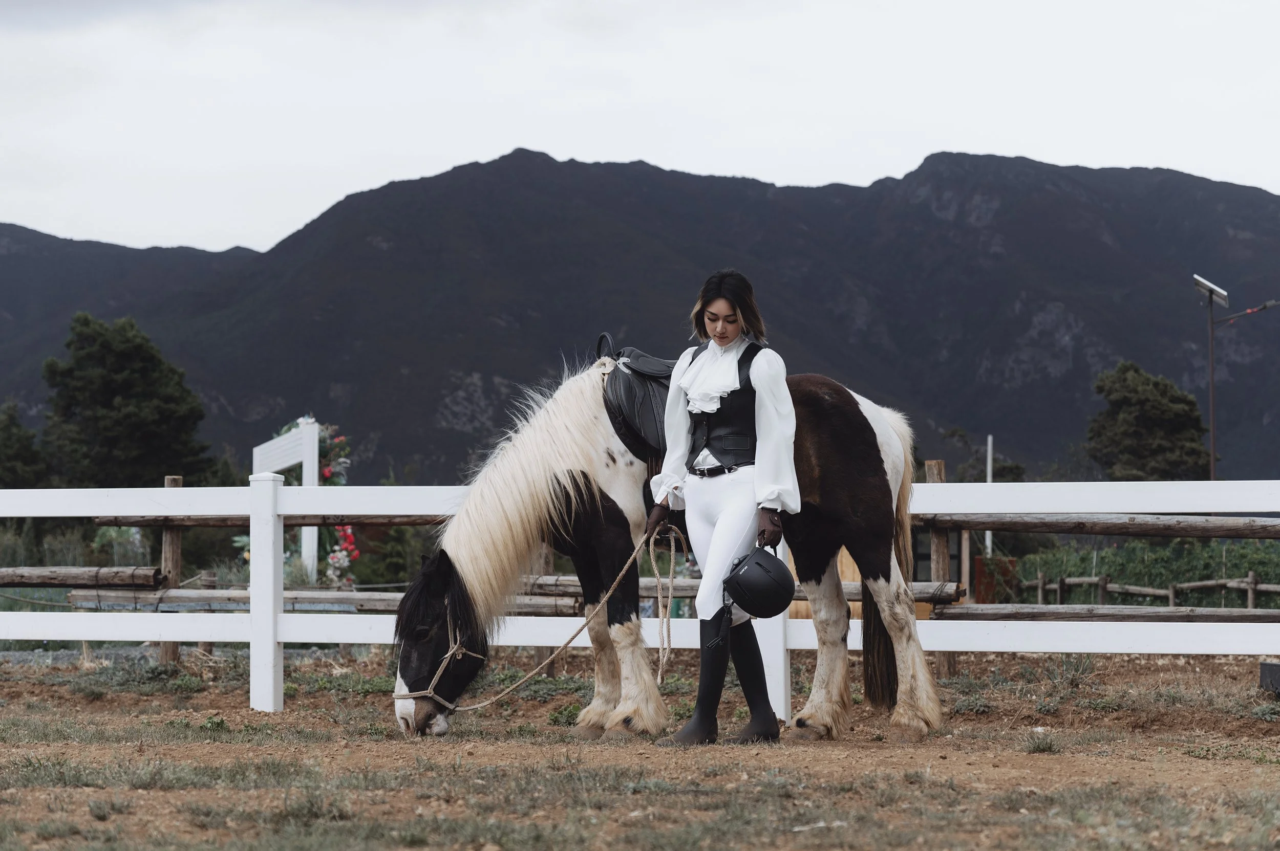 A woman dressed in white riding breeches and a white shirt, standing next to a horse in an outdoor riding arena. The horse is black and white, and the woman holds the reins while looking down at the horse. The background features mountains, trees, an