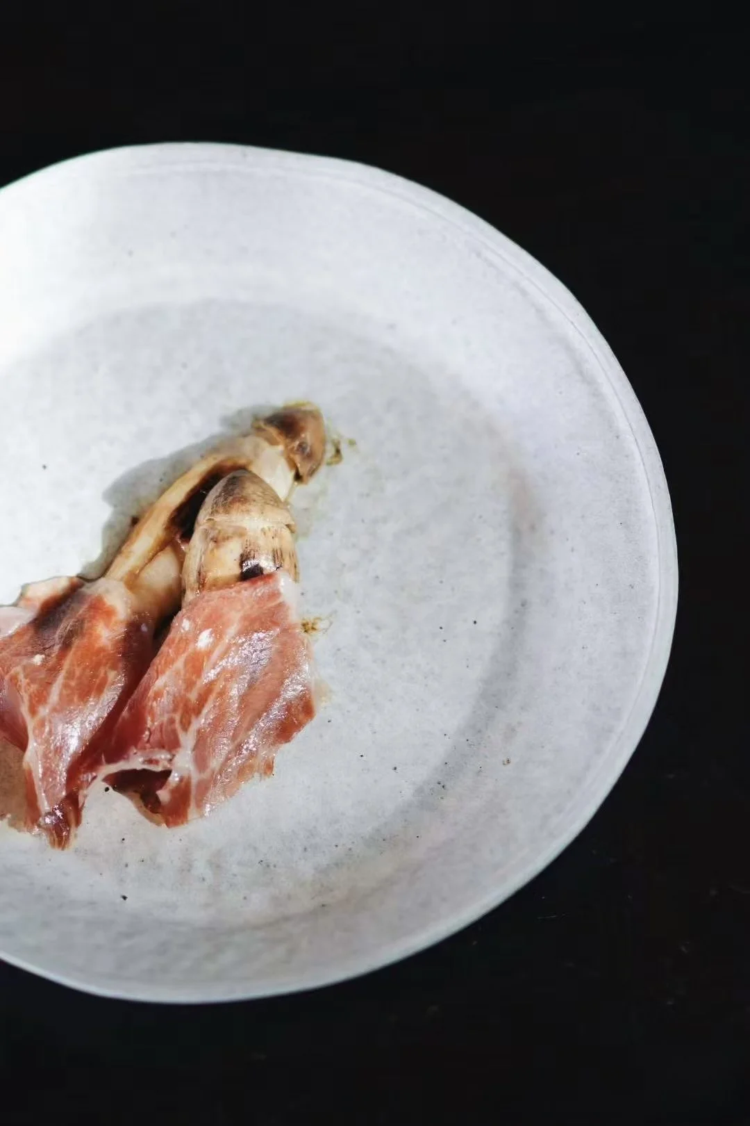 Close-up of a small plate with a piece of prosciutto-wrapped mushroom on a black background.