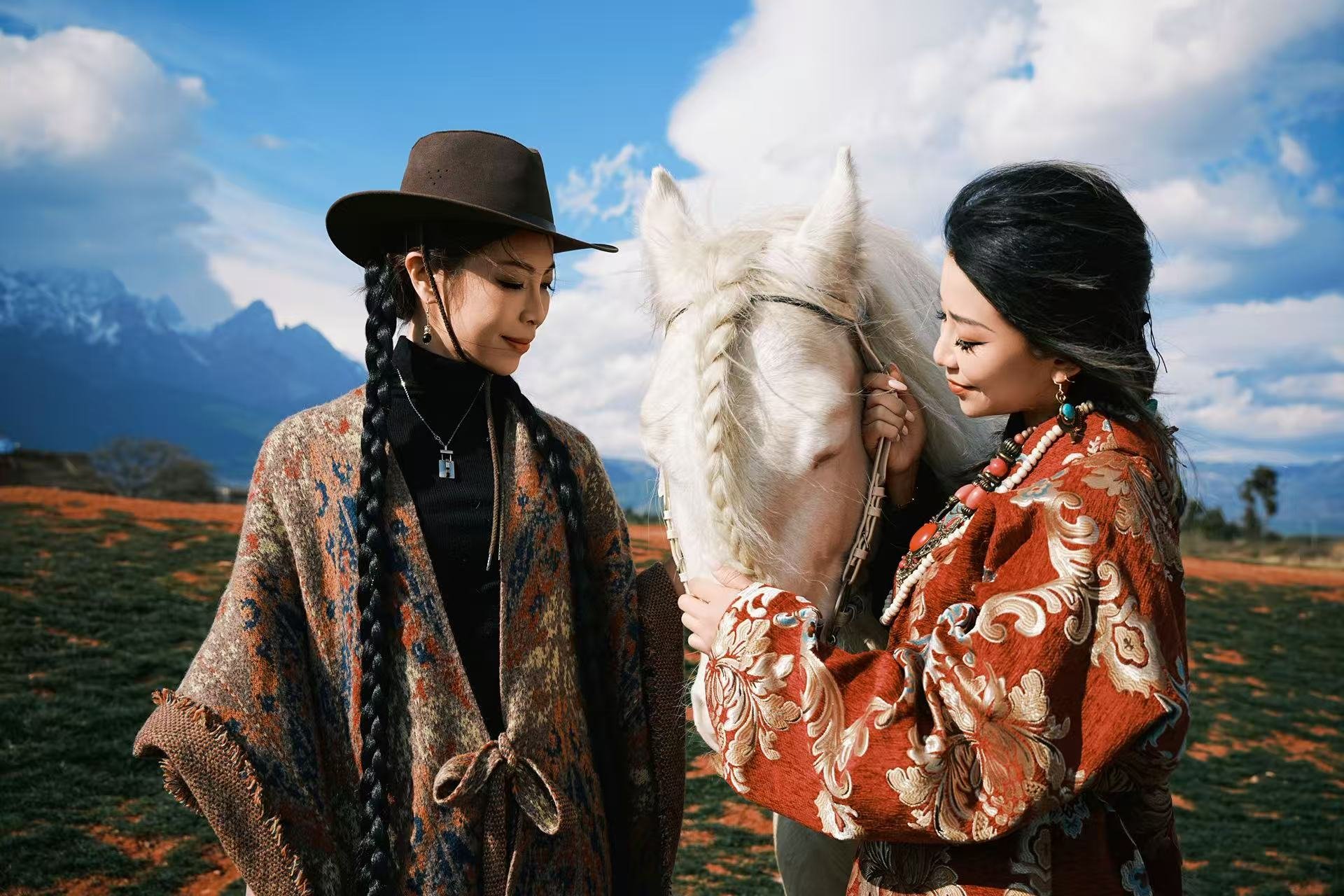 Two women with dark hair and patterned clothing gently touch a white horse's face outdoors with mountains and a partly cloudy sky in the background.