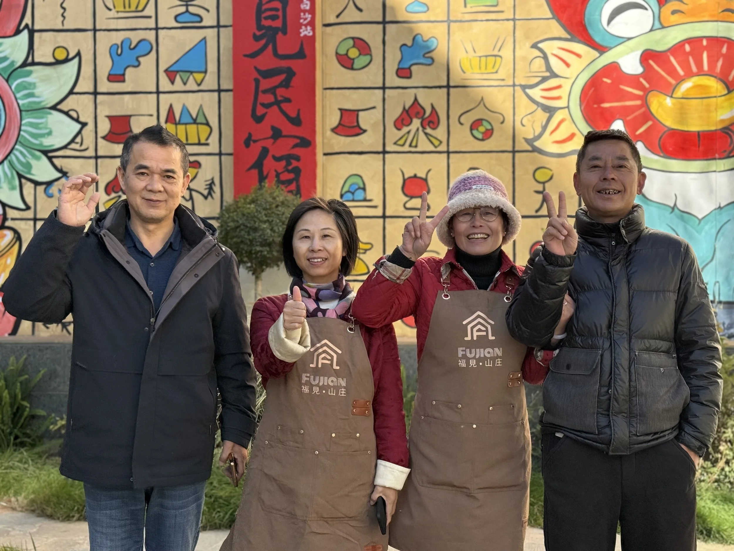 Four people standing outdoors in front of a colorful mural, smiling and making peace signs or thumbs-up gestures. The mural features bright, cartoon-style flowers, shapes, and Chinese characters.