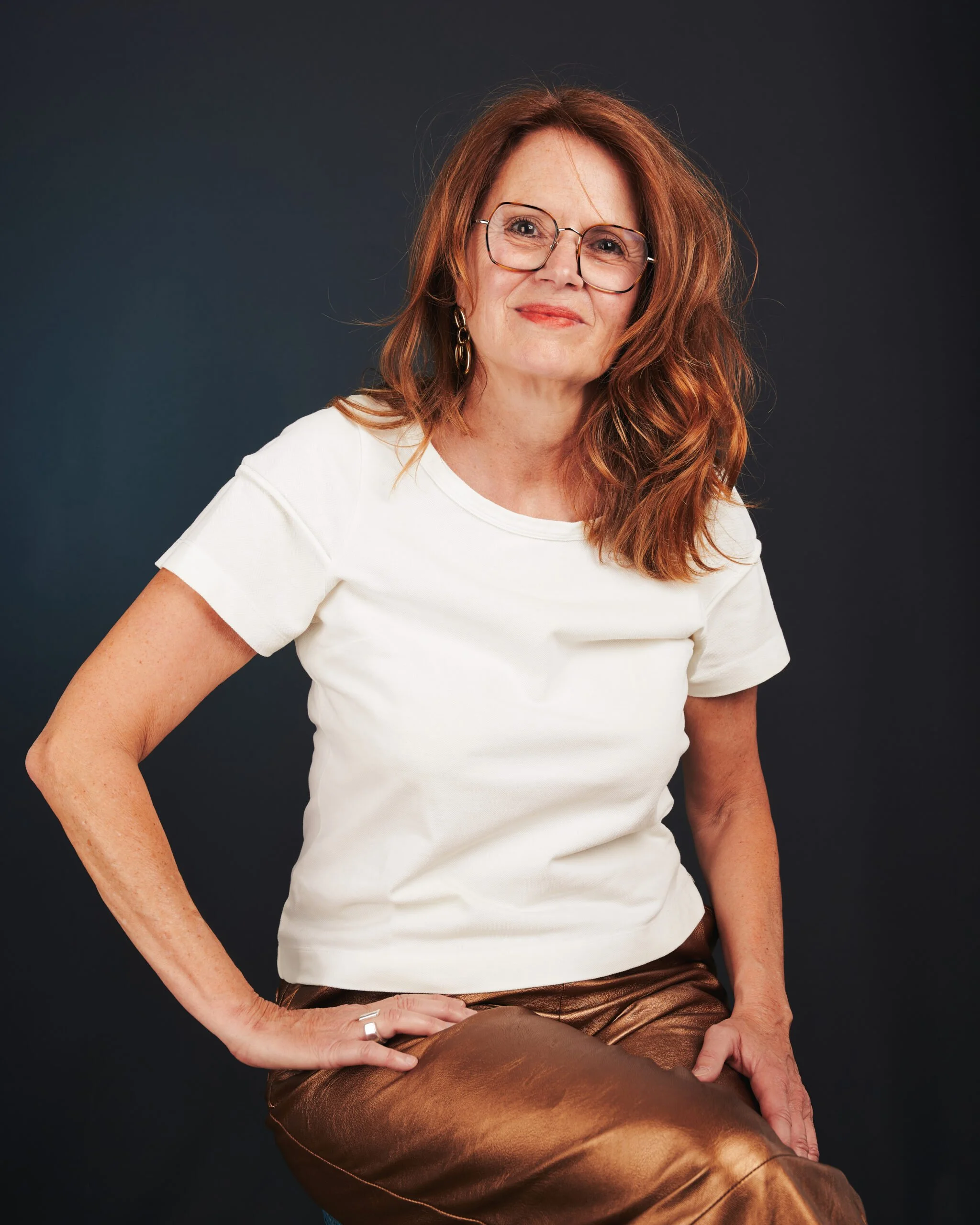 Une femme aux cheveux roux portant des lunettes, une chemise blanche et un pantalon en cuir marron, assise contre un fond gris foncé, souriant légèrement.
